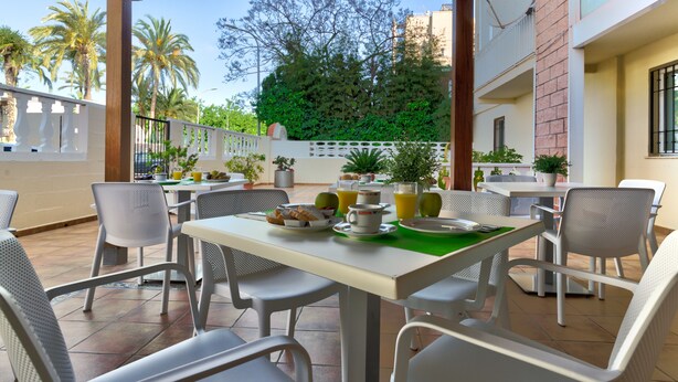 Un patio al aire libre con una mesa lista para el desayuno, rodeada de sillas y con vistas a palmeras y un edificio al fondo.