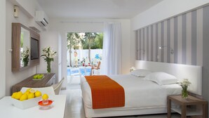 A modern hotel room with a bed, a bedside table, a desk with a fruit bowl, and a view of the outside. - Princessa Vera Hotel Apartments (Paphos)