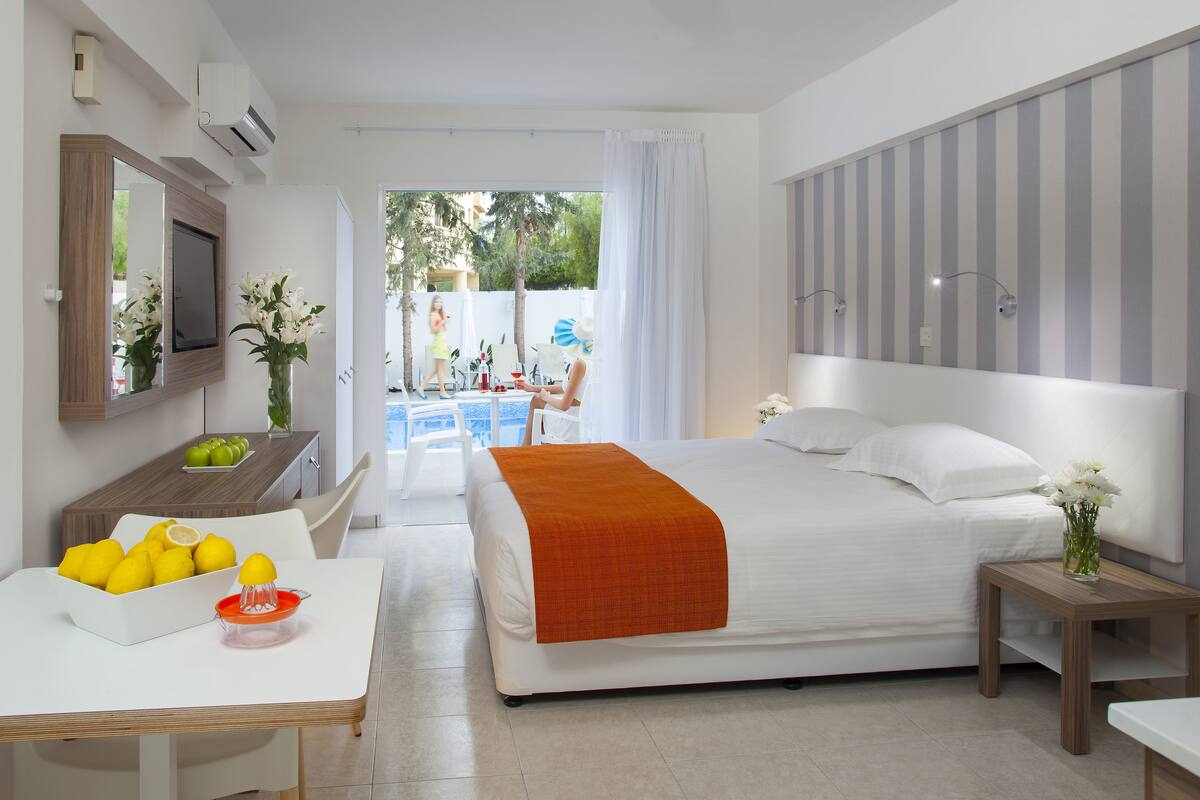 A modern hotel room with a bed, a bedside table, a desk with a fruit bowl, and a view of the outside.