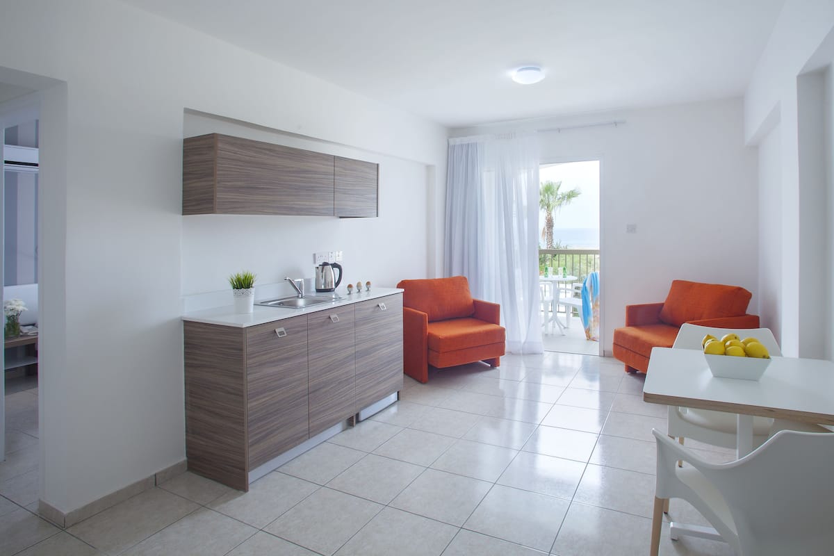 A modern kitchen with a wooden cabinet, a sink, and a coffee maker. There are two orange chairs and a white table with a bowl of lemons.