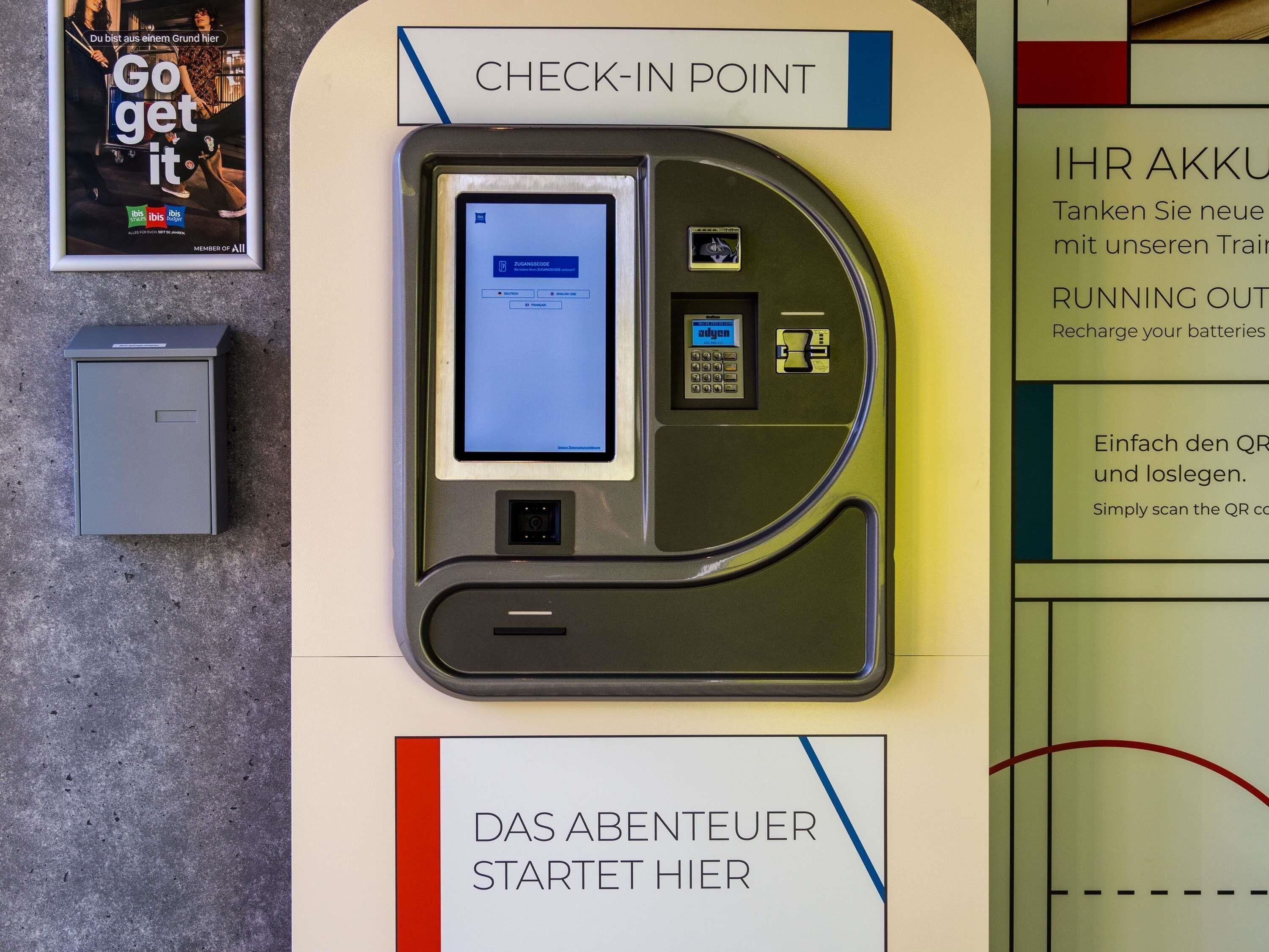 Un point d’enregistrement doté d’un terminal en libre-service, d’une petite armoire grise et d’un panneau avec du texte en allemand.