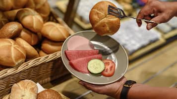 A person holding a plate with sliced meat, cucumber, and tomato, with a basket of bread in the background.