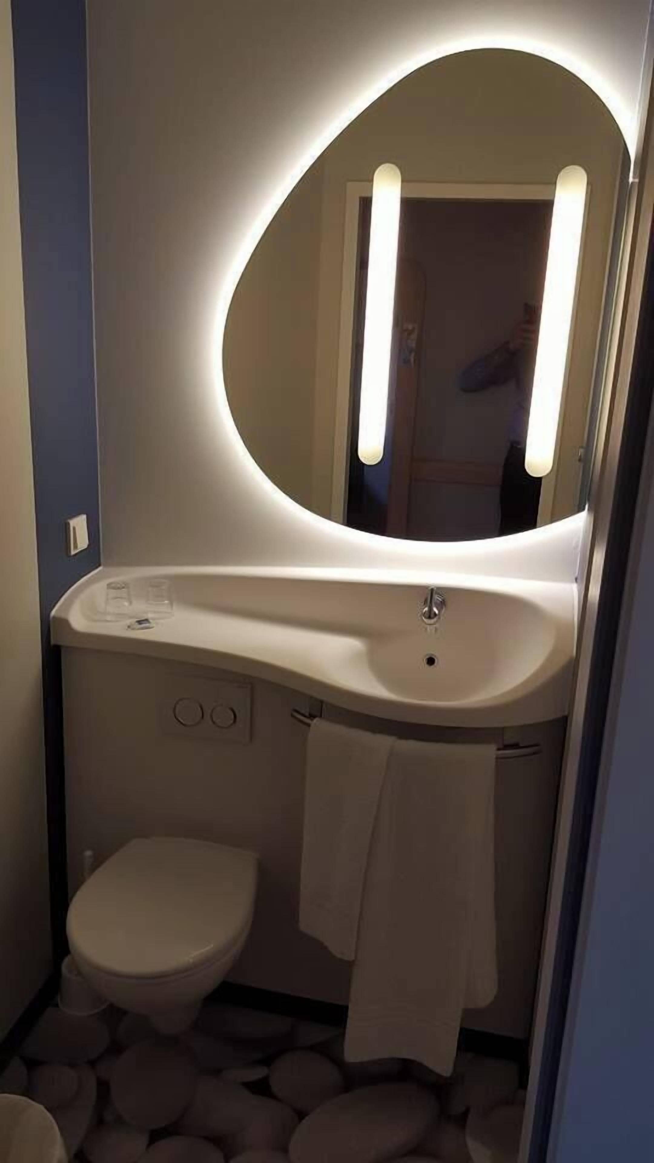 A bathroom with a white sink, a round mirror with lights, a toilet, and a towel hanging on the wall.
