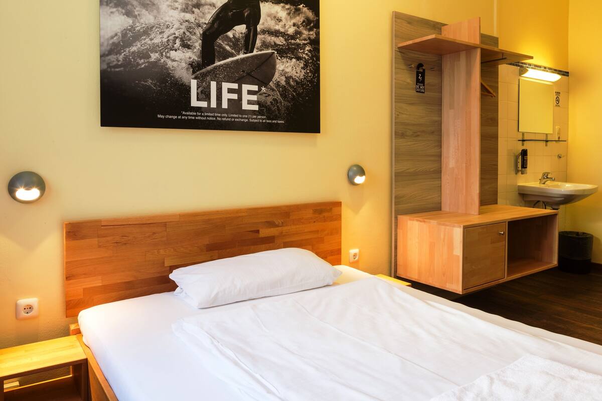 Une chambre d’hôtel avec une tête de lit en bois, un lit avec du linge de lit blanc, une armoire en bois, un miroir et une affiche accrochée au mur.