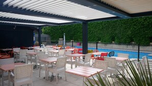 Un espacio al aire libre con mesas y sillas, una piscina y un muro de seto verde.
