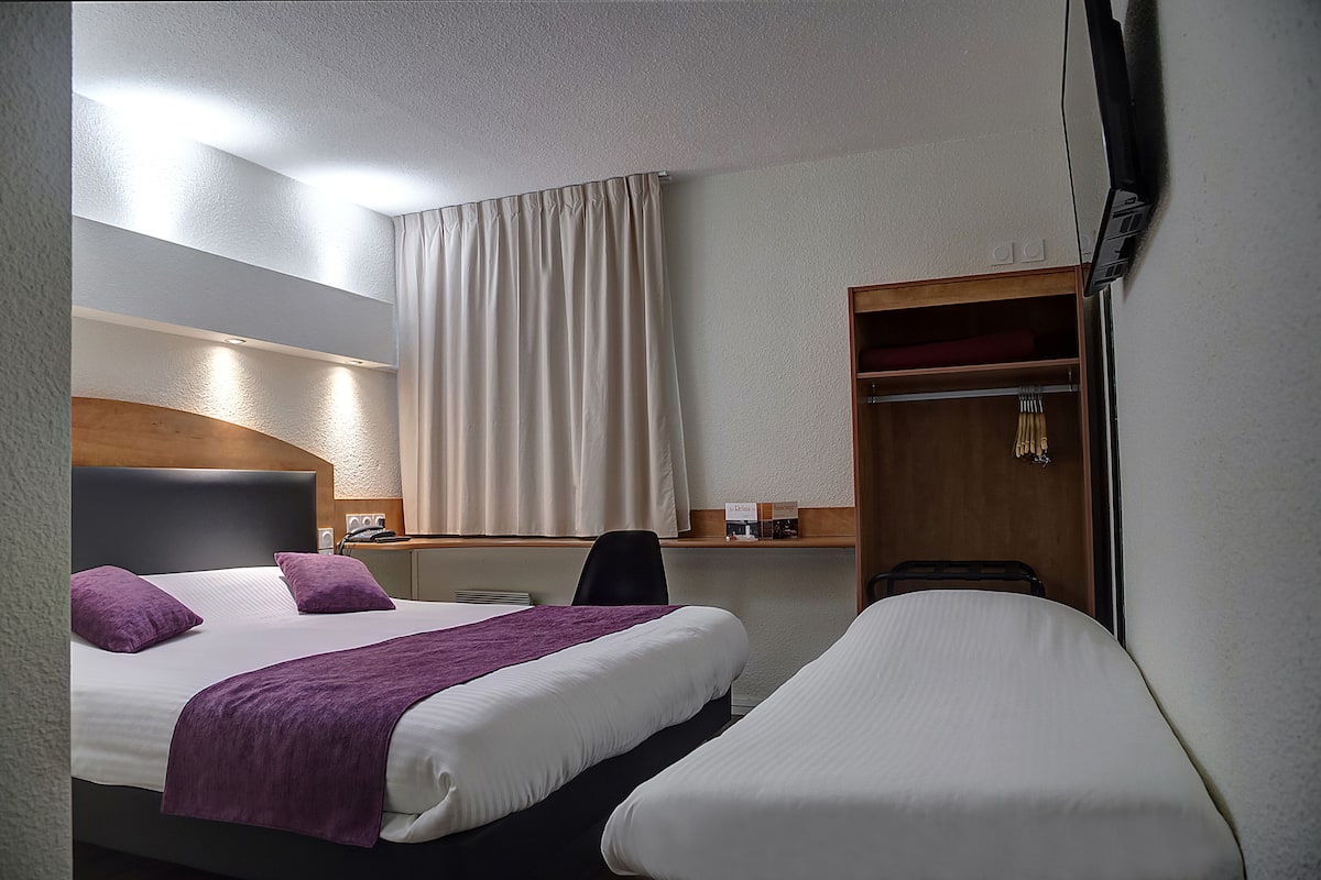 Una habitación de hotel con cama, escritorio, silla, armario y ventana con cortinas.