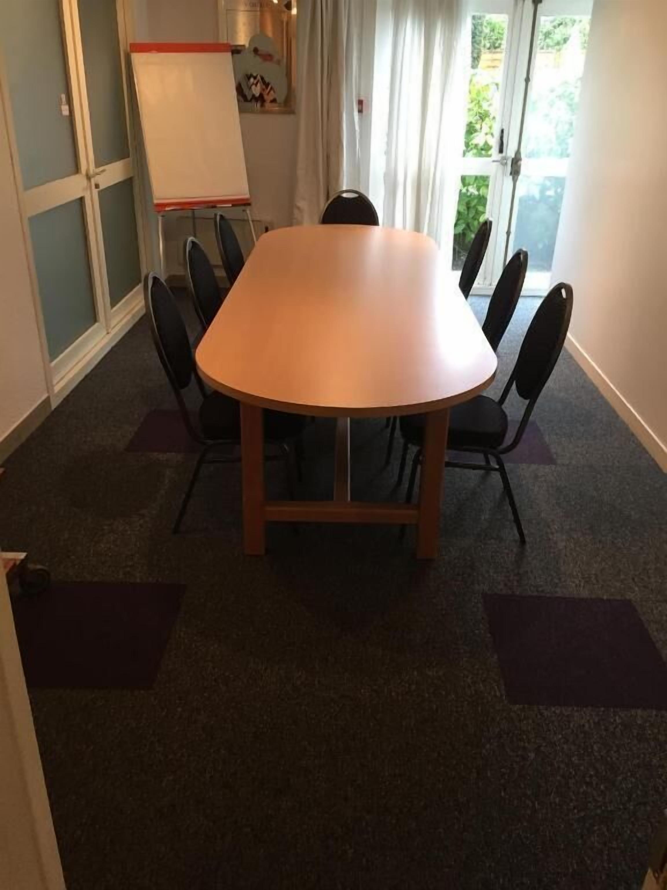A conference room with a large oval table, six chairs, a whiteboard, and a window with curtains.
