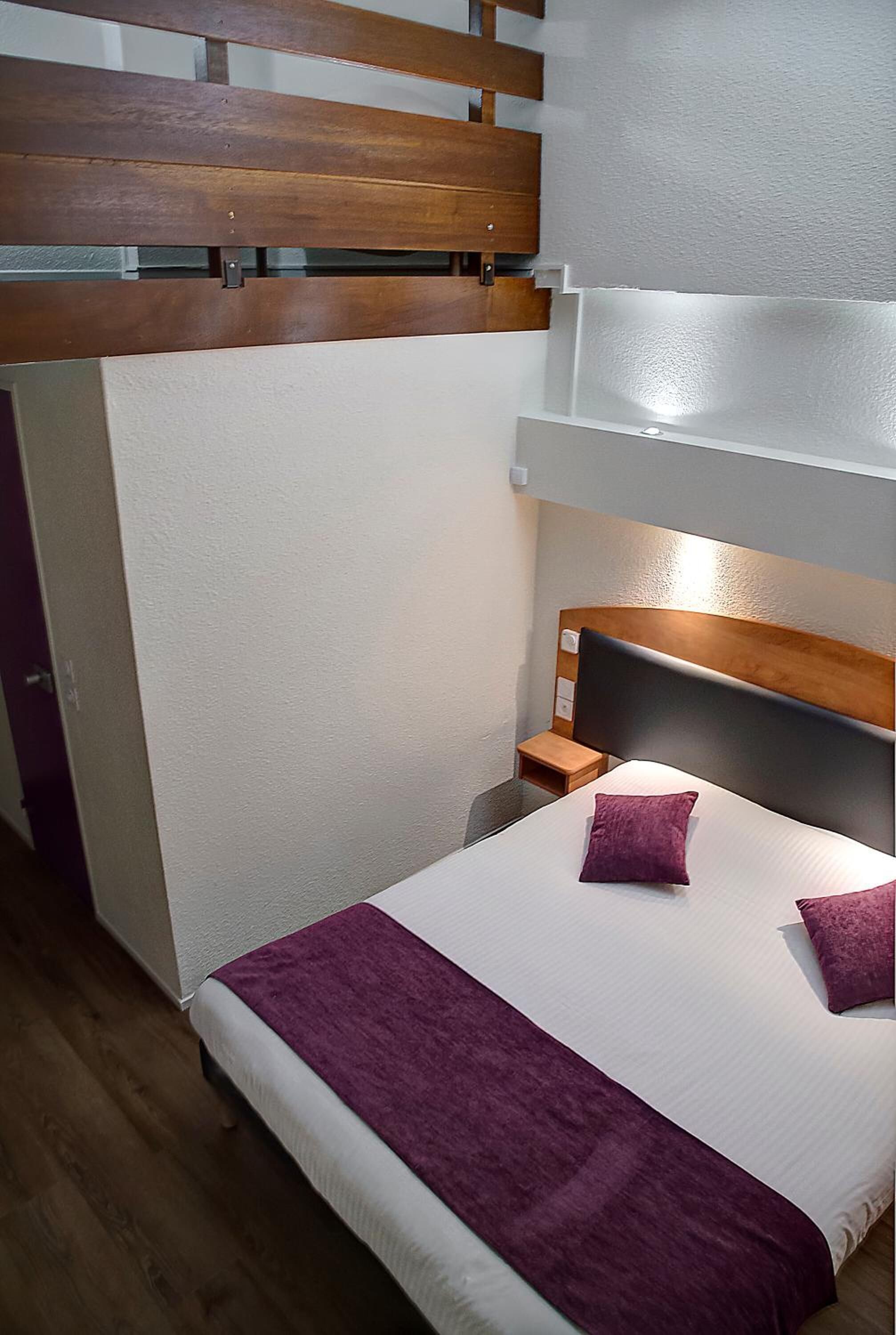 Habitación de hotel con cama, cabecera de madera, mesita de noche y una manta morada.