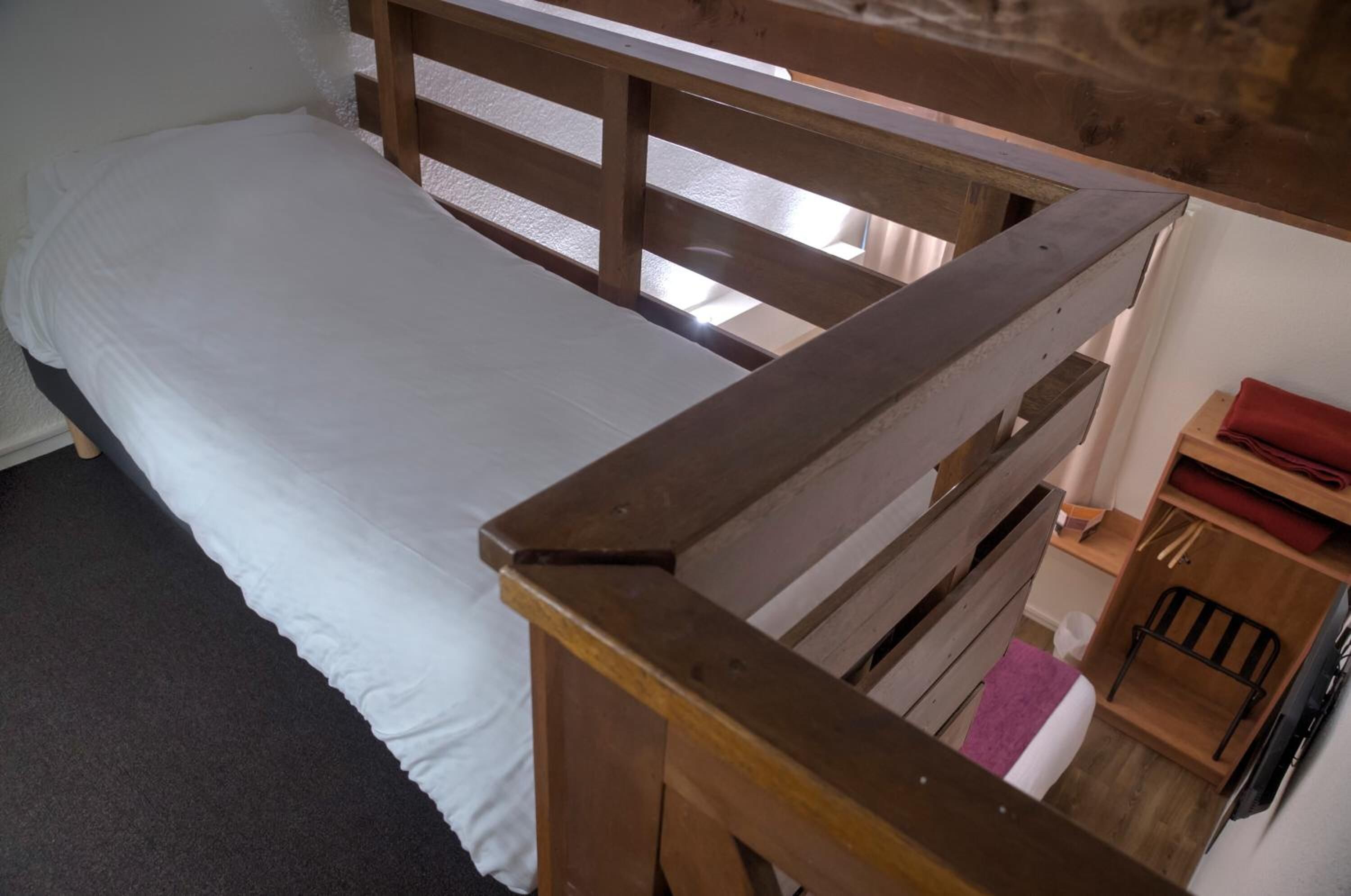Un lit simple avec une tête de lit en bois et un matelas blanc. Une petite étagère en bois avec un objet rouge est visible sur la droite.