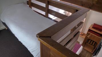 Un lit simple avec une tête de lit en bois et un matelas blanc. Une petite étagère en bois avec un objet rouge est visible sur la droite.