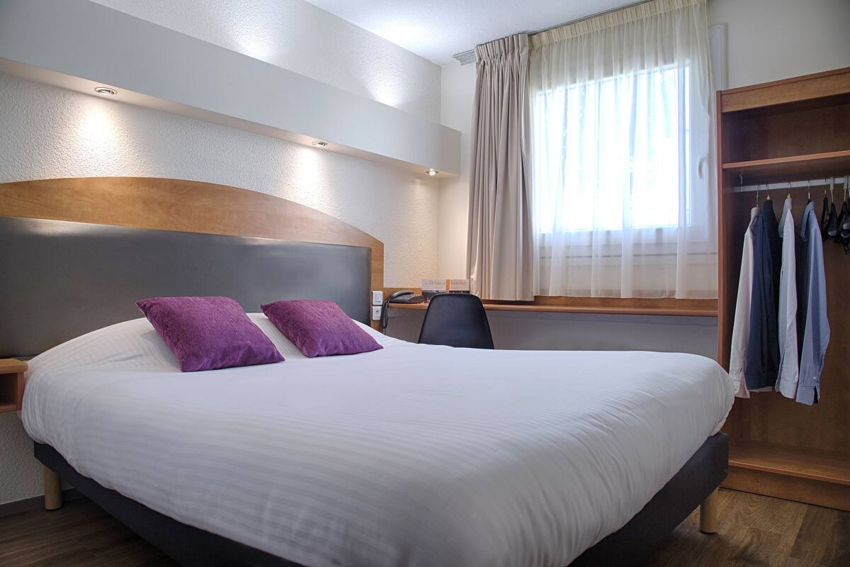 Habitación de hotel con una cama grande, cabecera de madera, un escritorio con silla y un armario con ropa colgada.