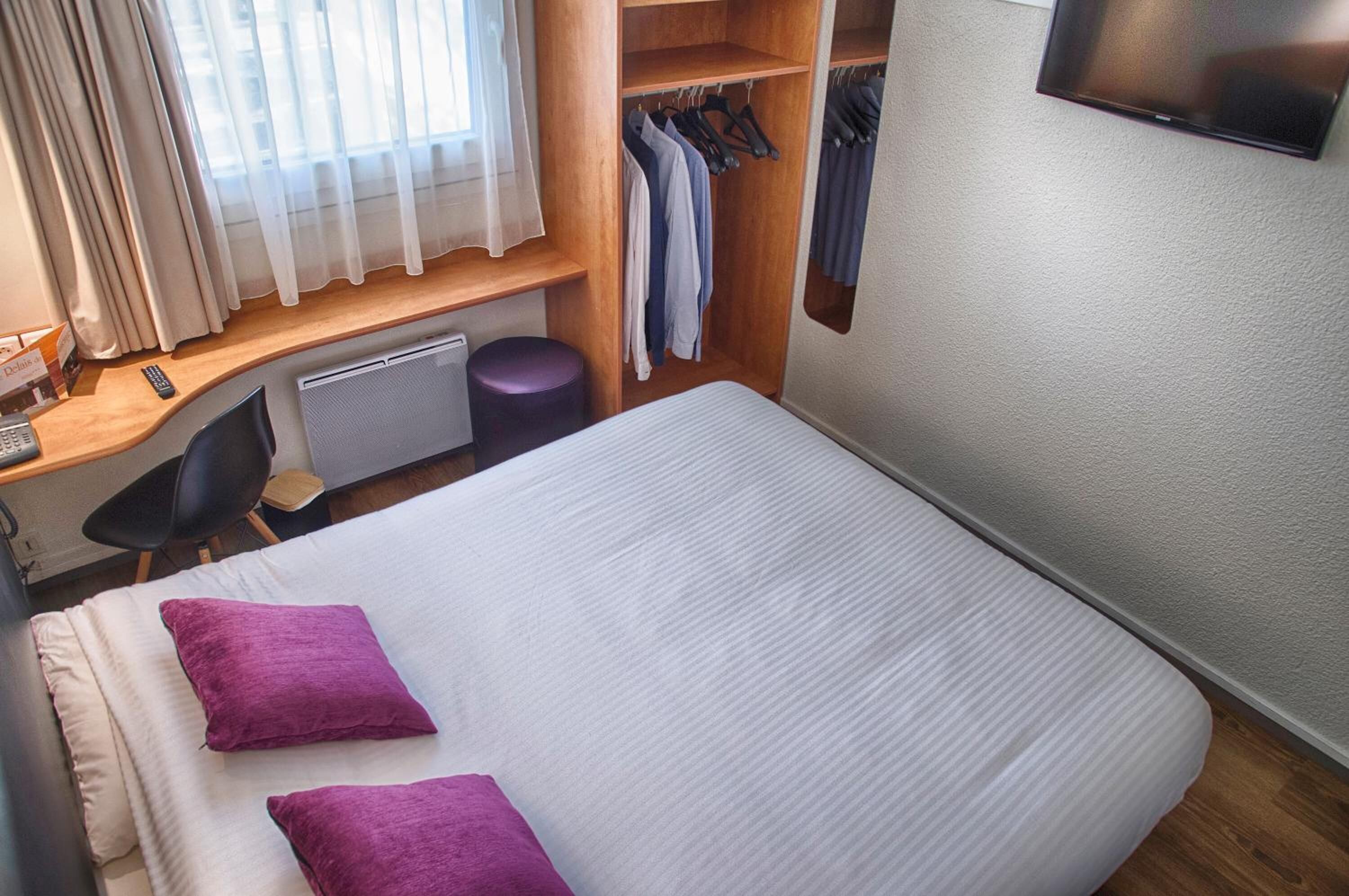 Habitación de hotel con cama, escritorio con silla, armario y televisor montado en la pared.
