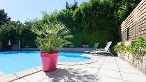 Una piscina con cubierta de baldosas, una planta rosada y dos sillas reclinables blancas.
