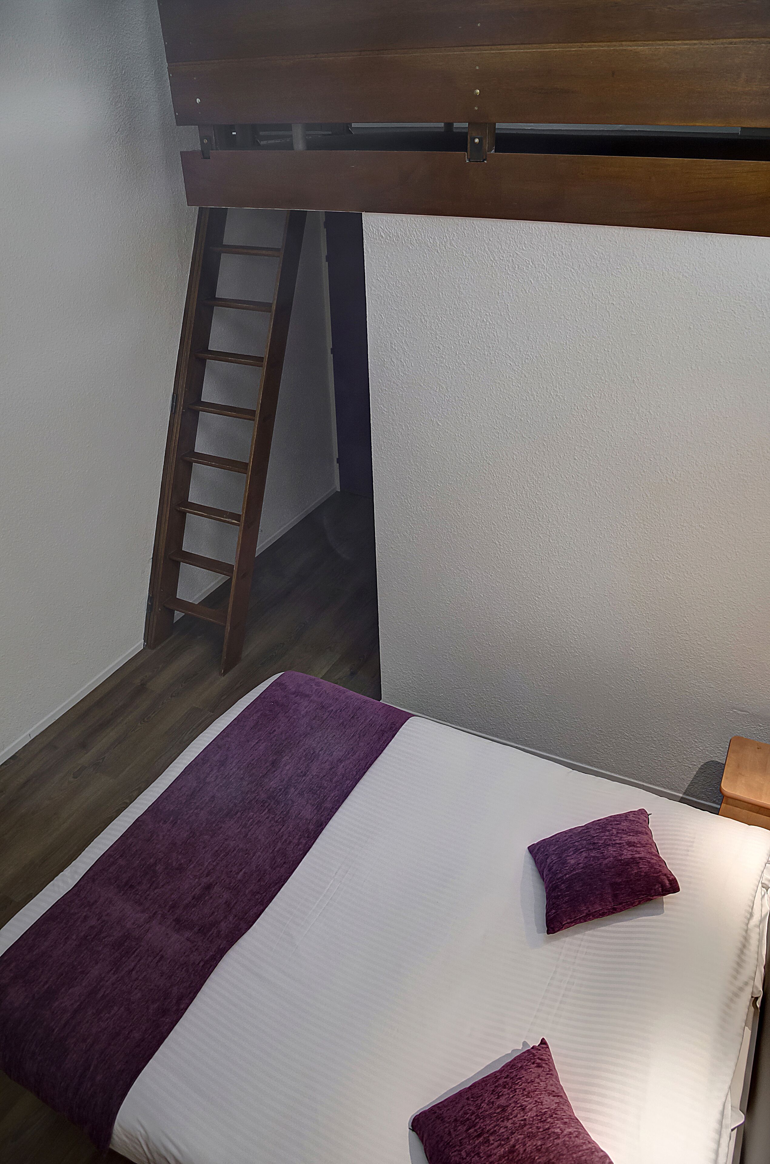Un dormitorio con una cama, una manta púrpura y una escalera de madera apoyada en la pared.