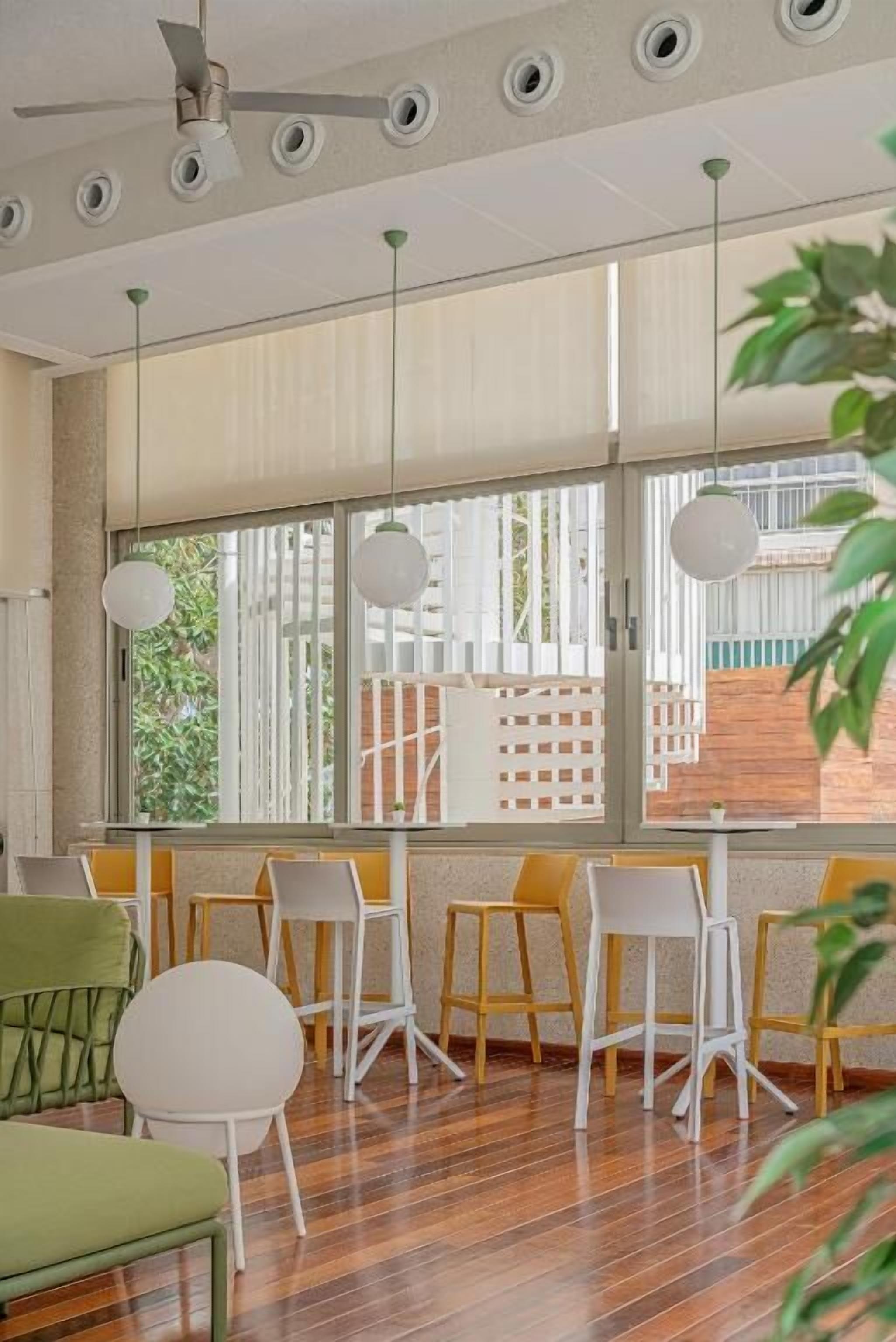 A modern room with a wooden floor, white and yellow chairs, a ceiling fan, and large windows with a view of greenery and a fence.