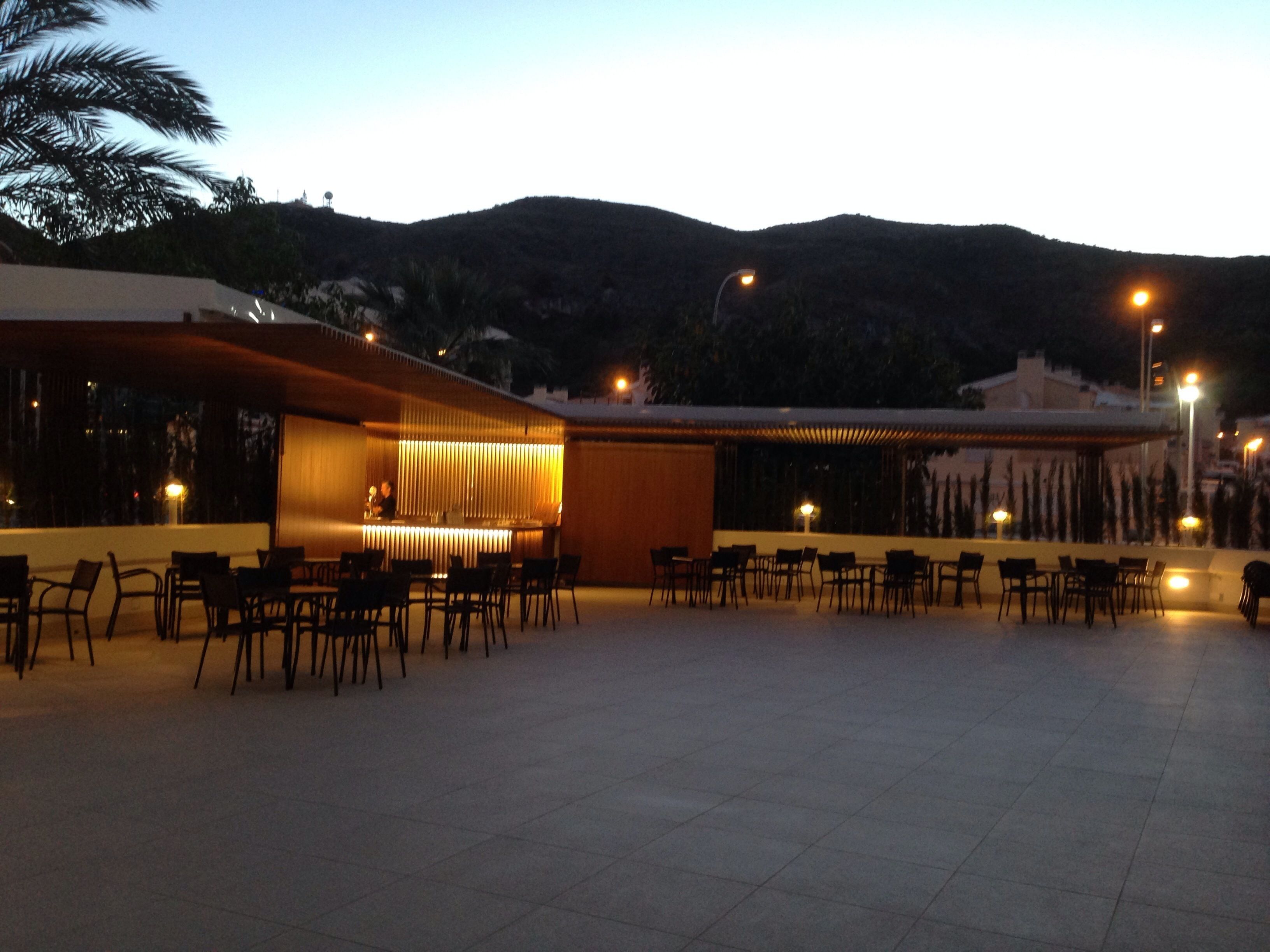 Un área de comedor al aire libre con varias mesas y sillas, un bar con mostrador y taburetes, y vistas a un paisaje montañoso al fondo.