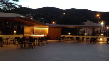 Un área de comedor al aire libre con varias mesas y sillas, un bar con mostrador y taburetes, y vistas a un paisaje montañoso al fondo.