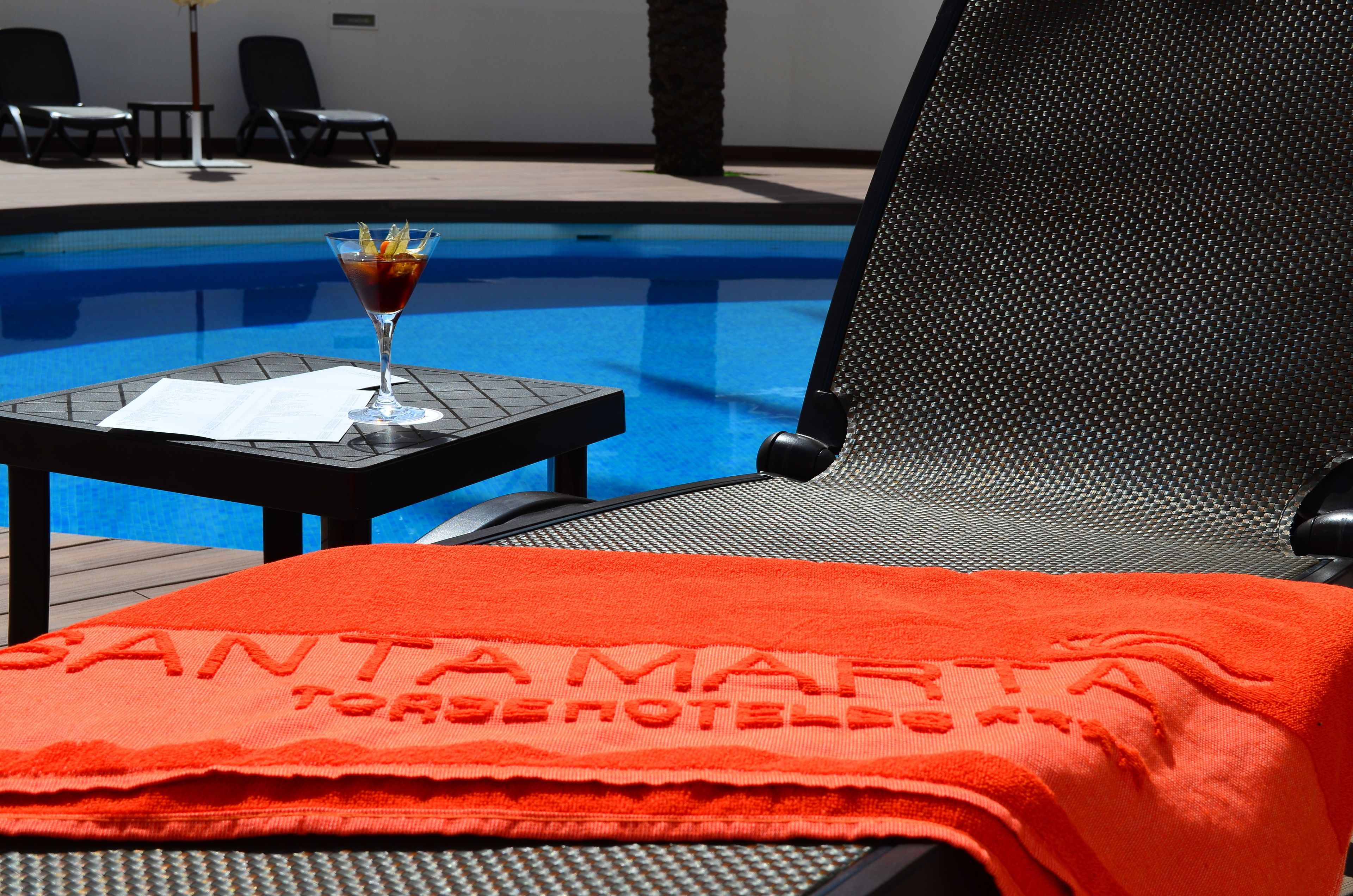 A poolside area with a lounge chair, a table with a drink, and a towel with text.