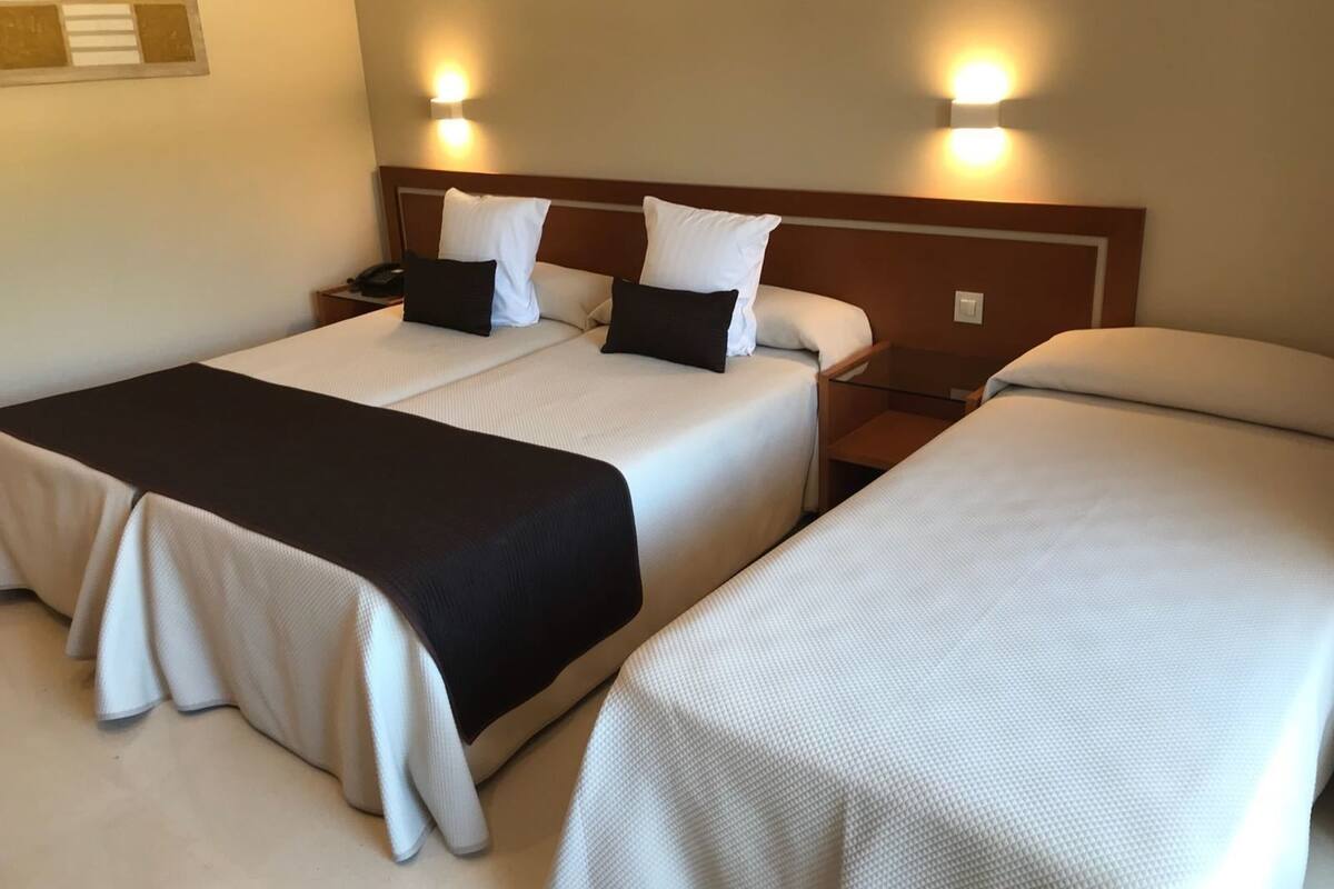 A hotel room with two beds, each with white bedding and black throw pillows. There are bedside tables with lamps and a wall-mounted light fixture.