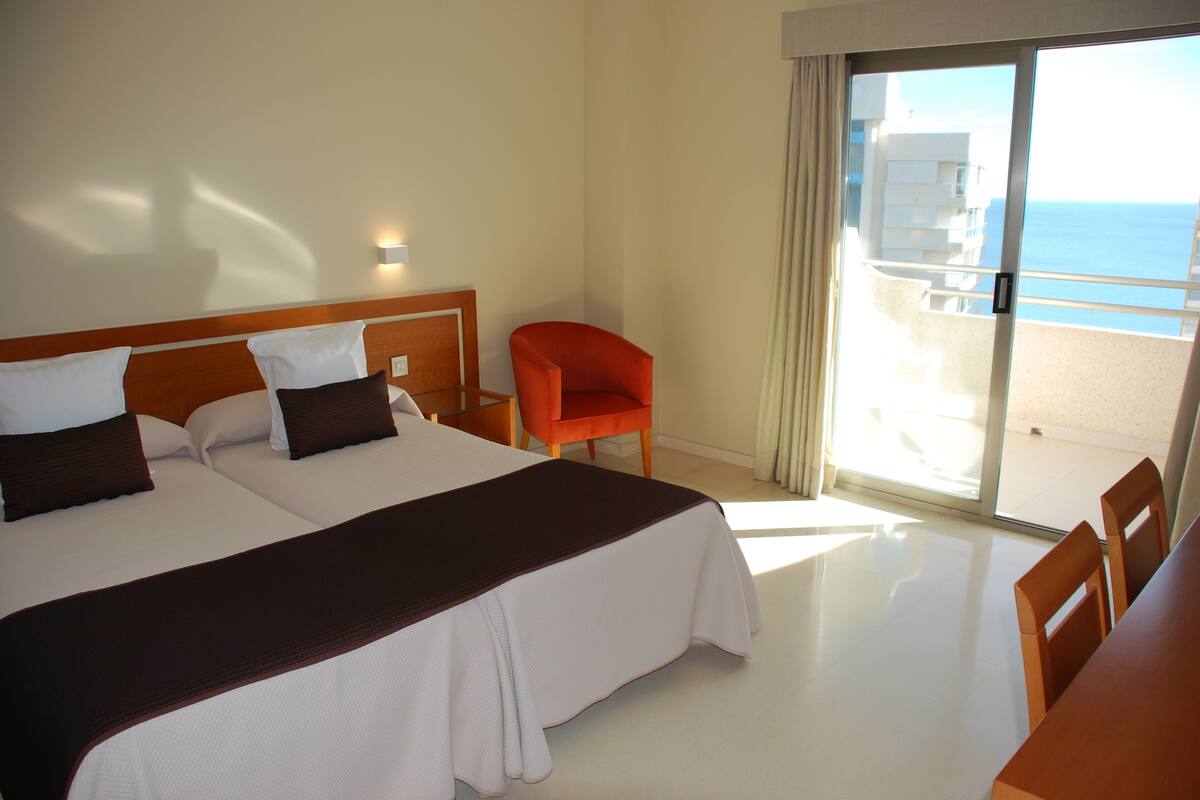 A hotel room with two beds, a chair, and a balcony with a view of the sea.
