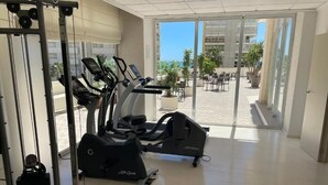 A gym with cardio machines and a weight rack, large windows, and a view of outdoor seating and palm trees.
