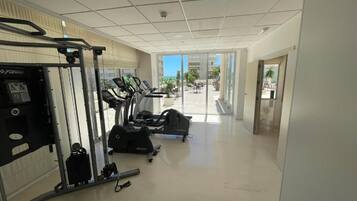 Un gimnasio con máquinas de cardio, equipo de levantamiento de pesas y vista despejada al exterior, con palmeras y un edificio al fondo.
