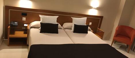 A double bed with two black pillows, a wooden nightstand with a phone, and a red chair.