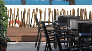 An outdoor seating area with wicker chairs and tables, a wooden fence, and a view of the ocean and palm trees.
