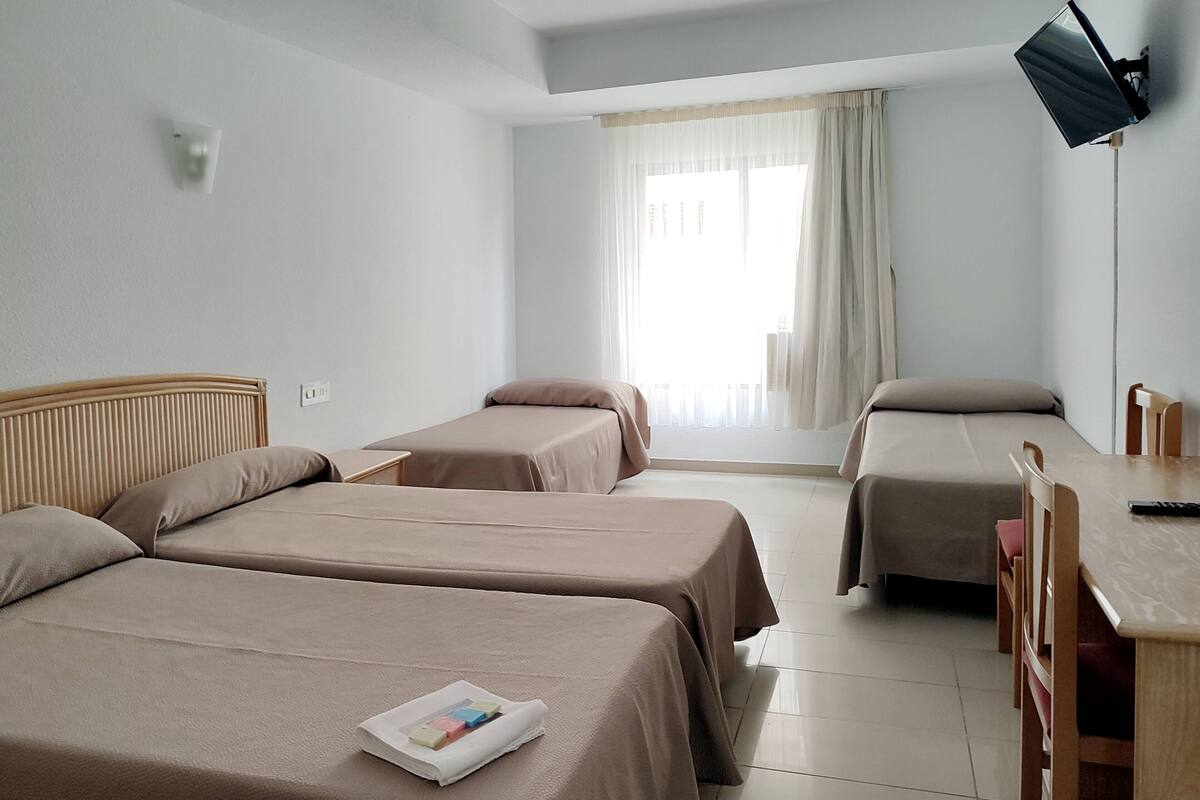 Quarto de hotel com duas camas de solteiro, cabeceira de madeira, escrivaninha com cadeira, TV de tela plana na parede e janela com cortinas.