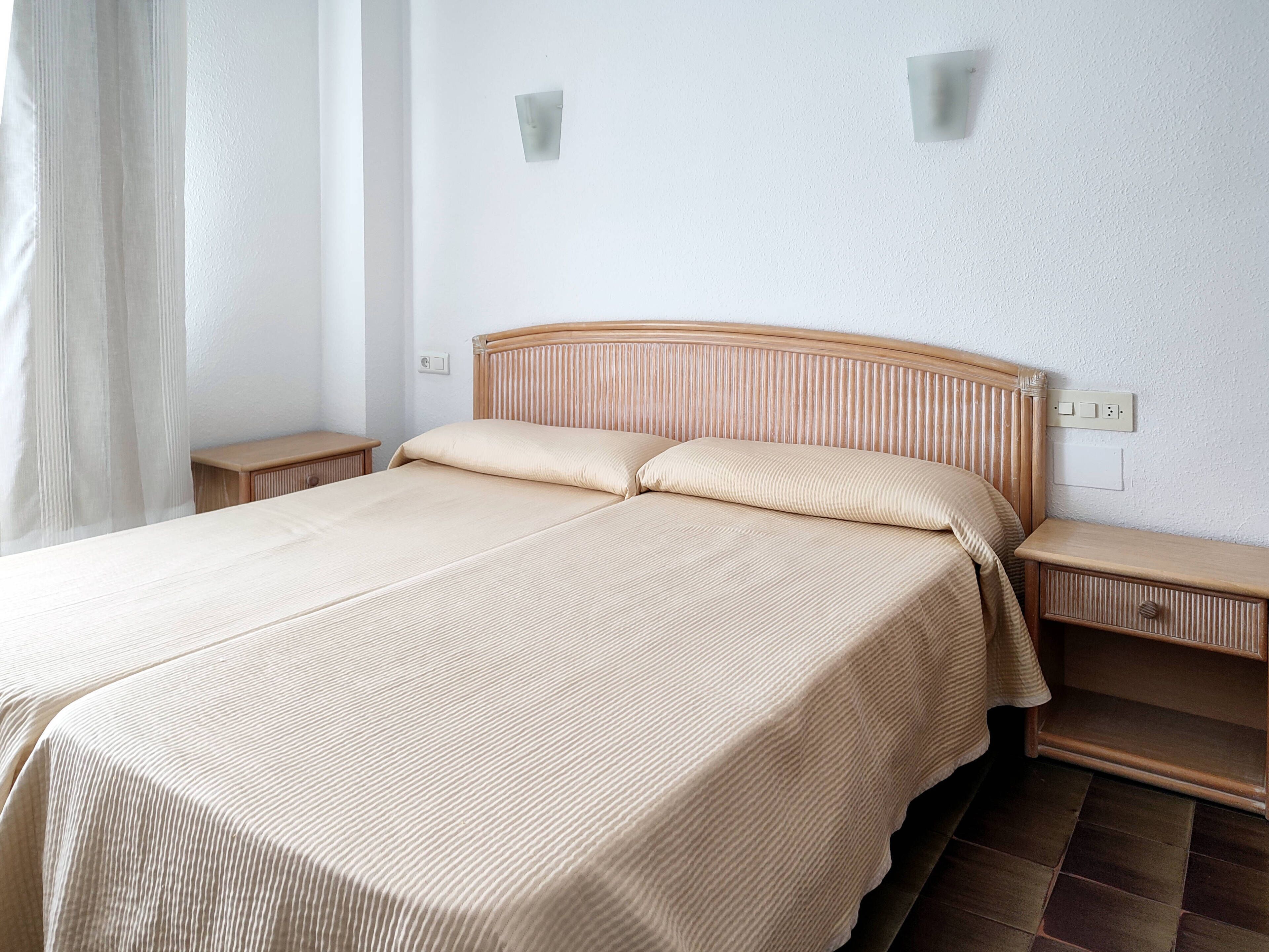 Una cama bien hecha con cabecera de madera, dos mesitas de noche y una pared blanca lisa con un enchufe.