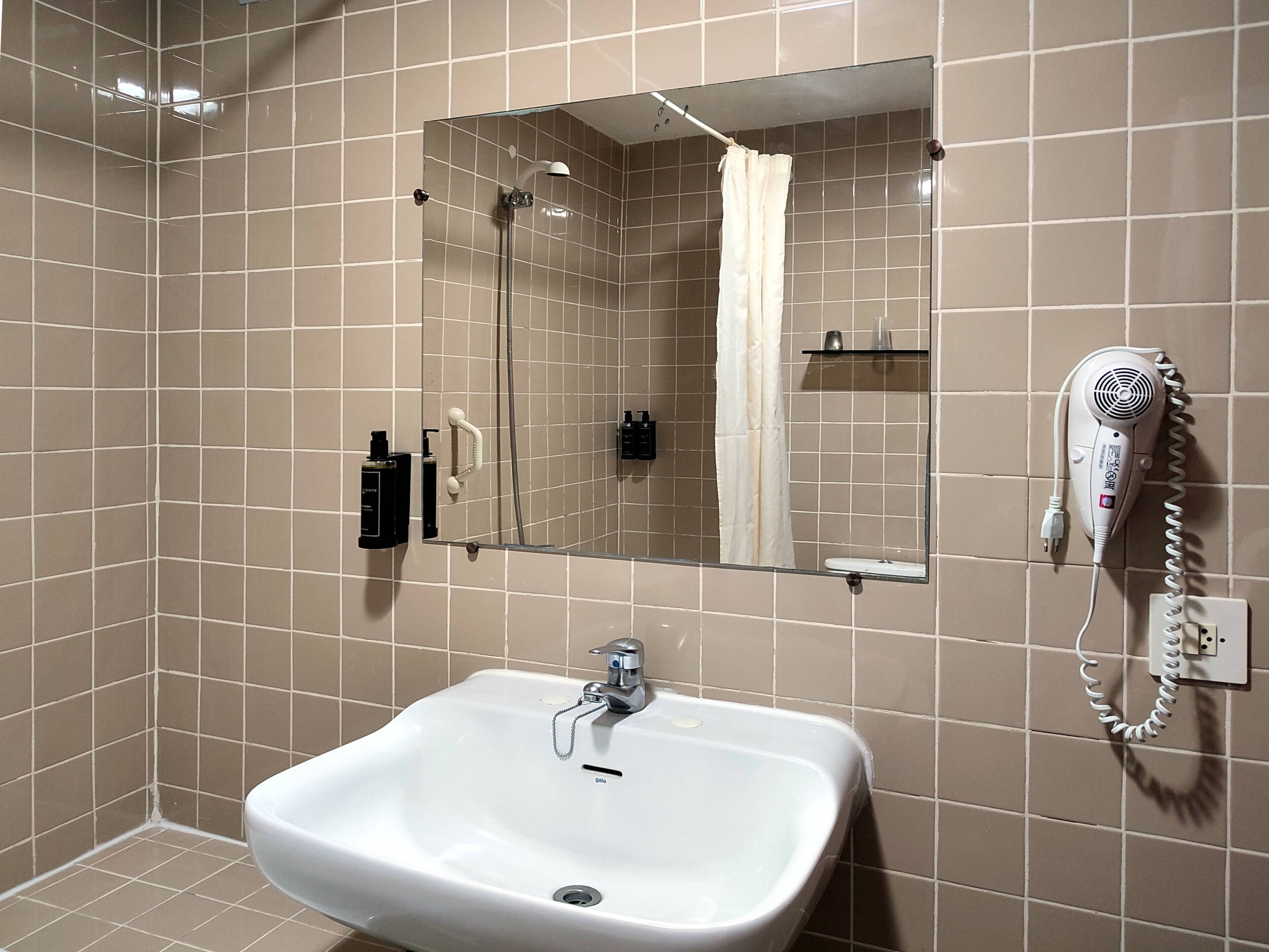 Un baño con un lavamanos blanco, un espejo, una ducha y un secador de pelo montado en la pared.