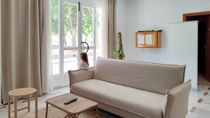 A living room with a beige sofa, a small wicker table with a remote control, a wicker chair, and a large window with curtains. - Hotel Porto (Gandia)