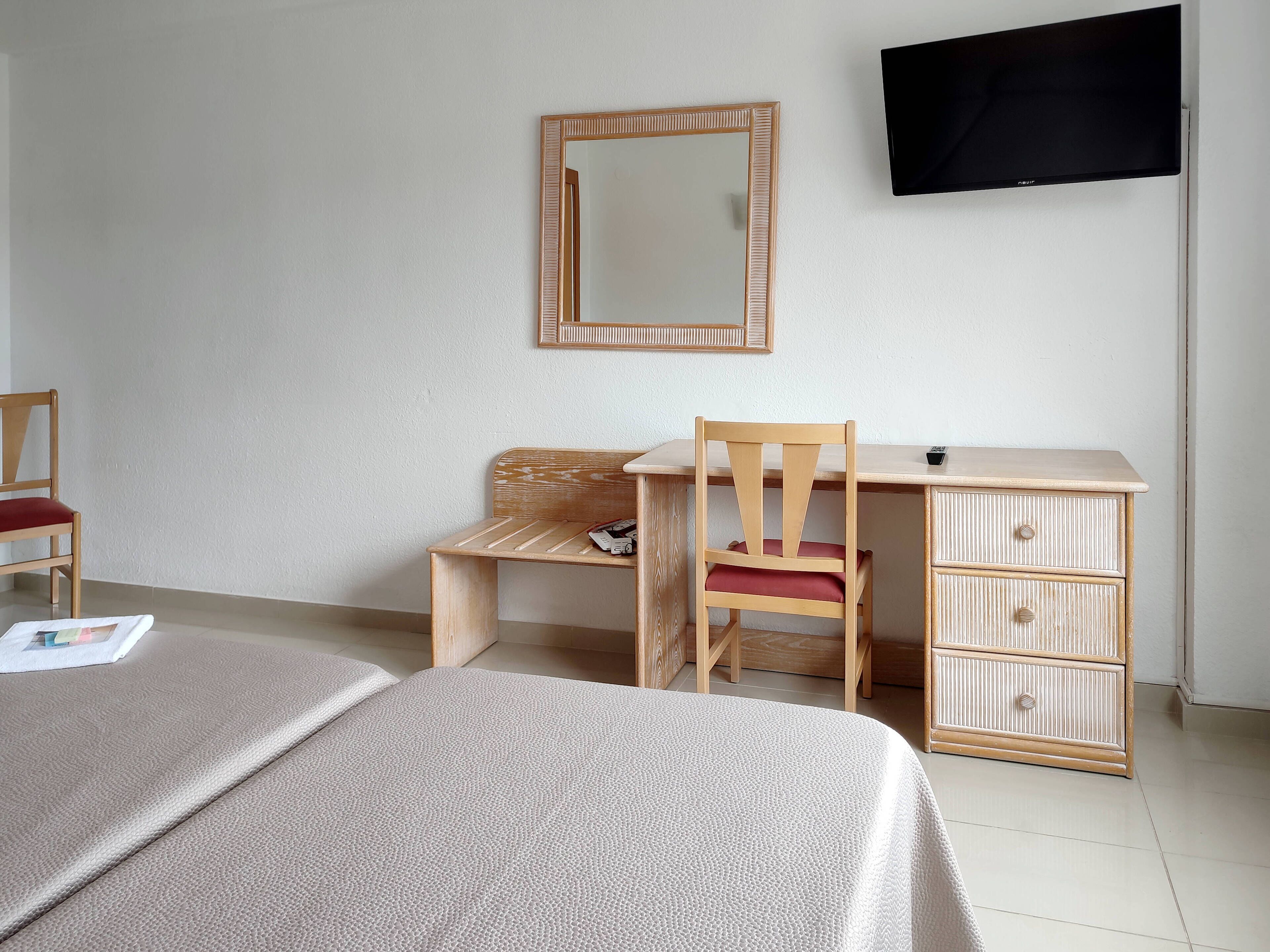 Una habitación con una cama, un escritorio de madera, una silla, un aparador y un televisor en la pared.