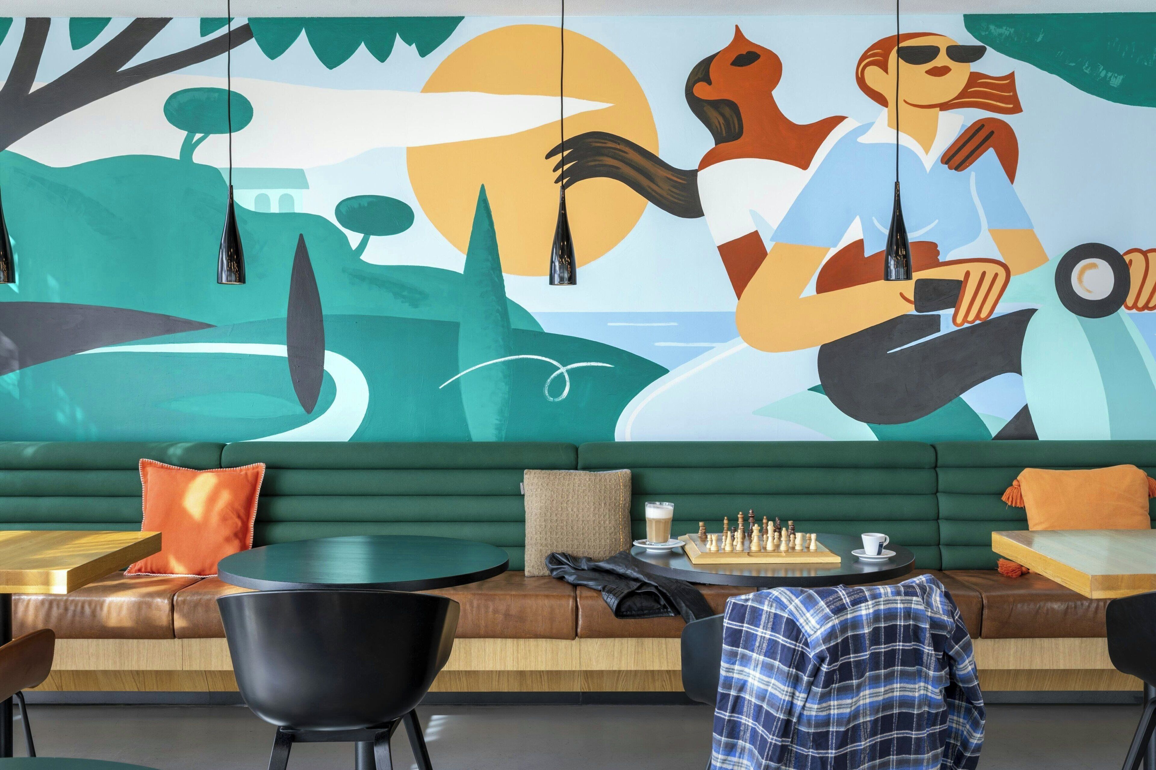 A modern lounge area with a green sofa, black chairs, a chess table, and a mural on the wall.