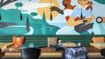 A modern lounge area with a green sofa, black chairs, a chess table, and a mural on the wall.