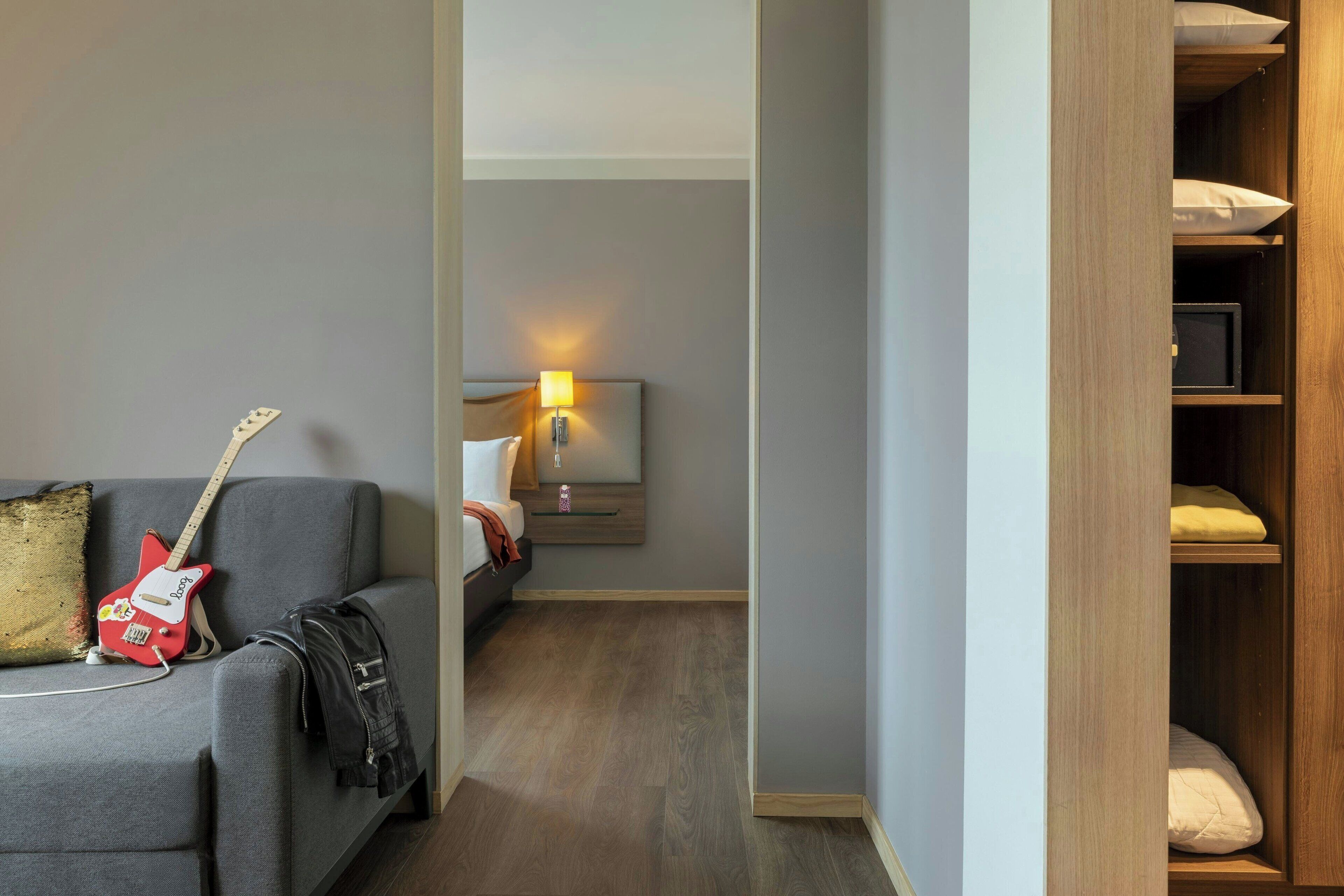 A hotel room with a grey sofa, a red electric guitar, a black jacket, a bedside table with a lamp, and a wooden wardrobe.