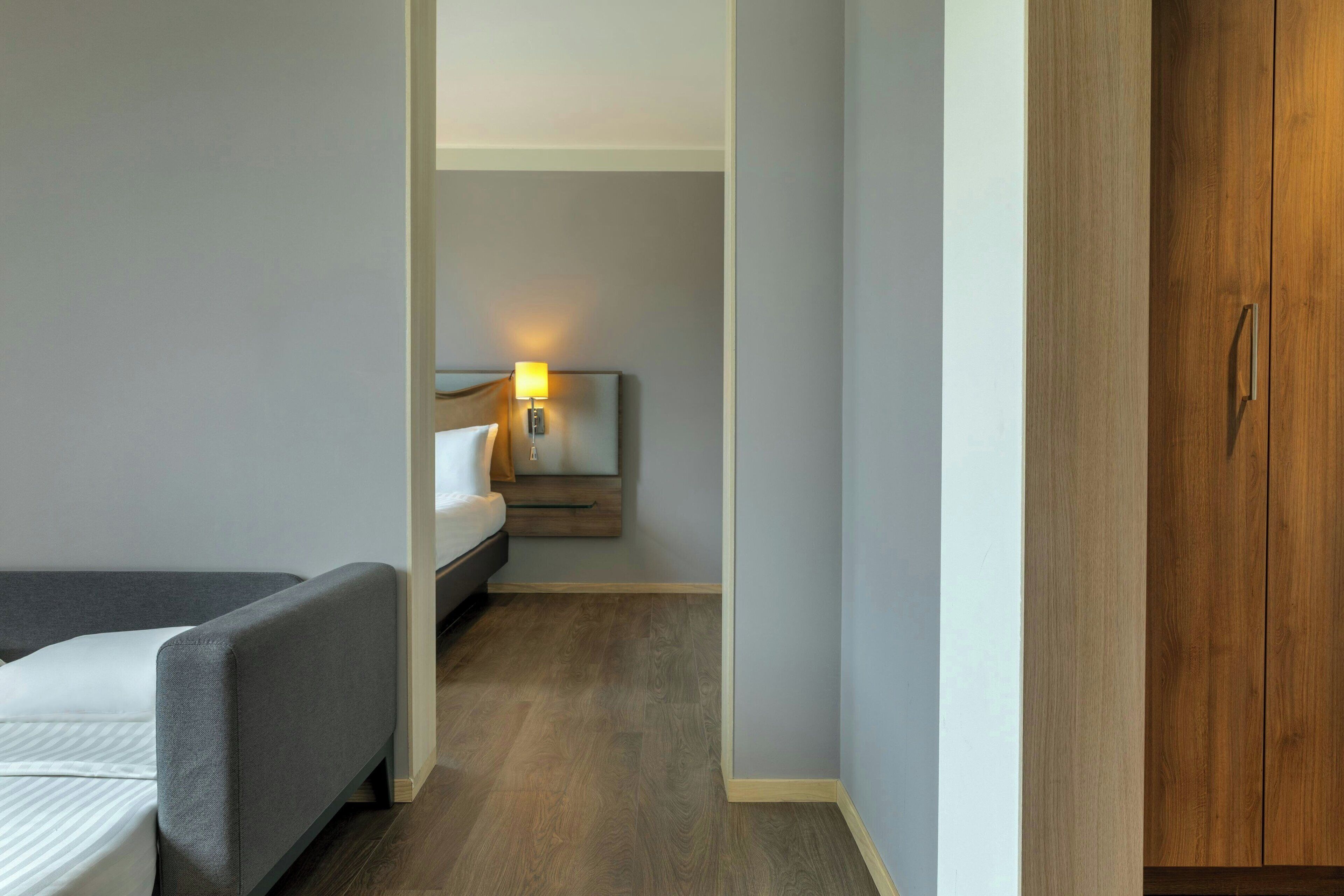 Een smalle gang die leidt naar een slaapkamer met een bed, nachtkastje en een lamp.