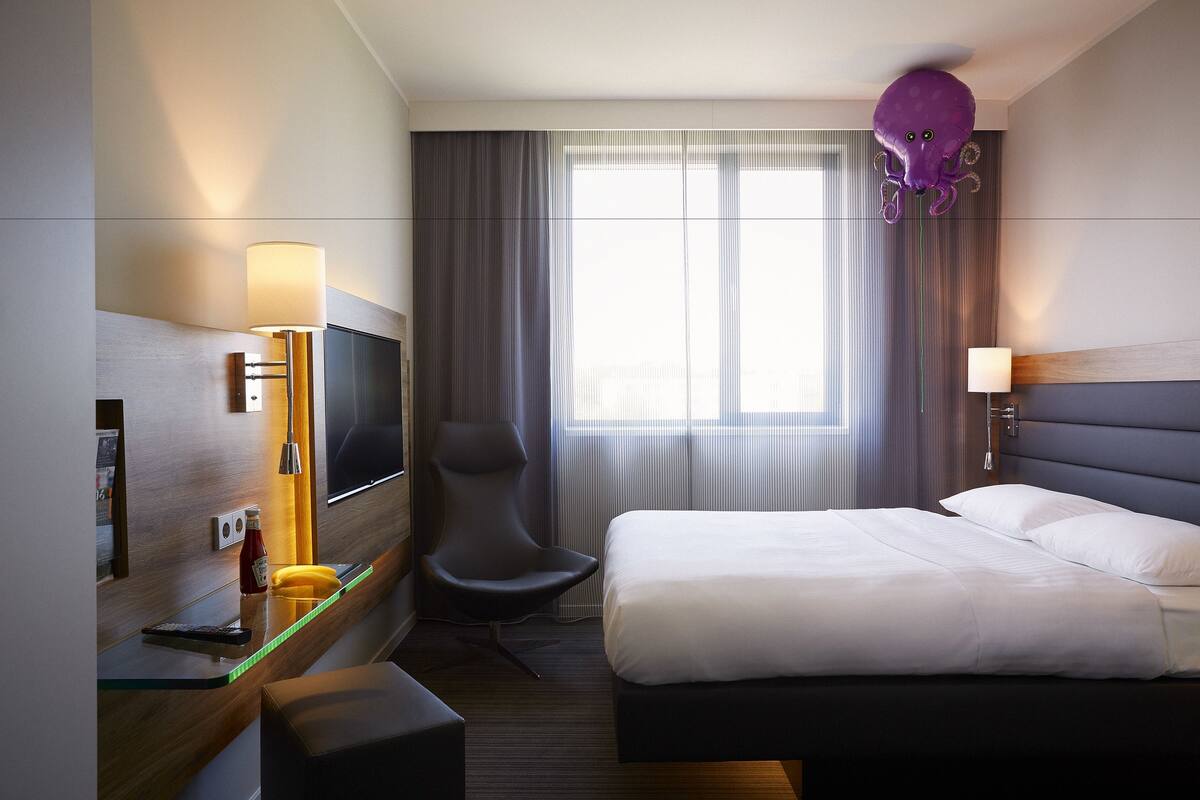 A hotel room with a bed, a desk with a lamp, a chair, a TV, and a window with curtains.