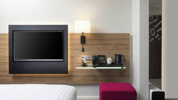 Habitación de hotel con televisor de pantalla plana montado en la pared, una mesita de noche con lámpara, cafetera y purificador de aire Subcool.