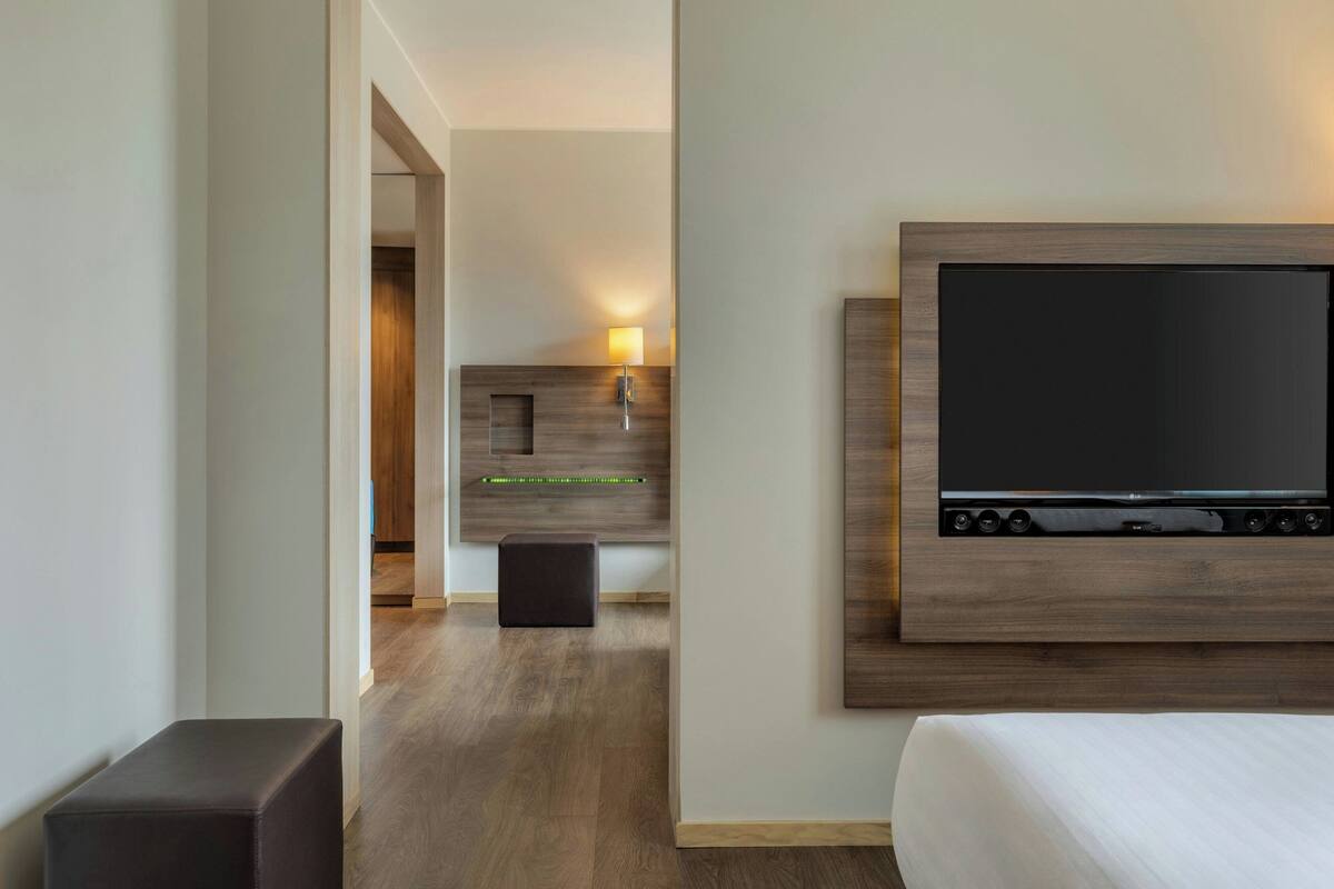 A modern hotel room with a large flat-screen TV mounted on the wall, a wooden bedside table, and a bench.