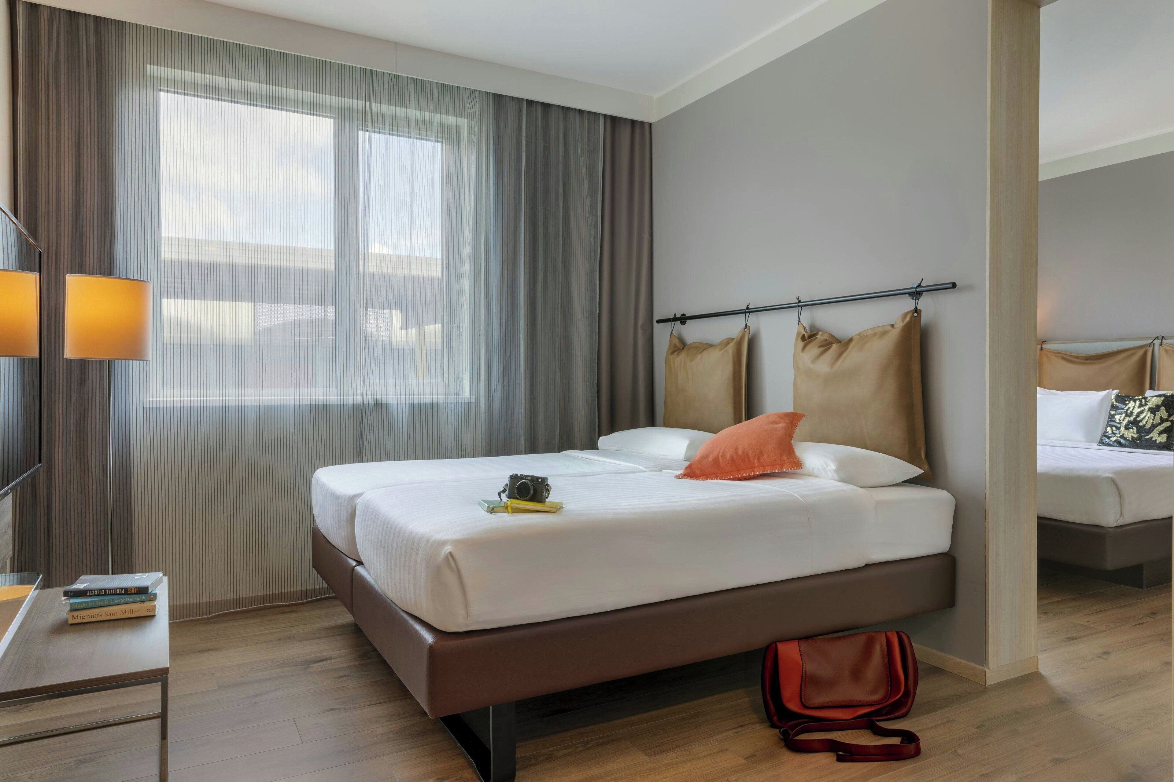 Habitación de hotel con una cama grande, una mesita de noche con lámpara, una ventana con cortinas y otra cama al fondo.