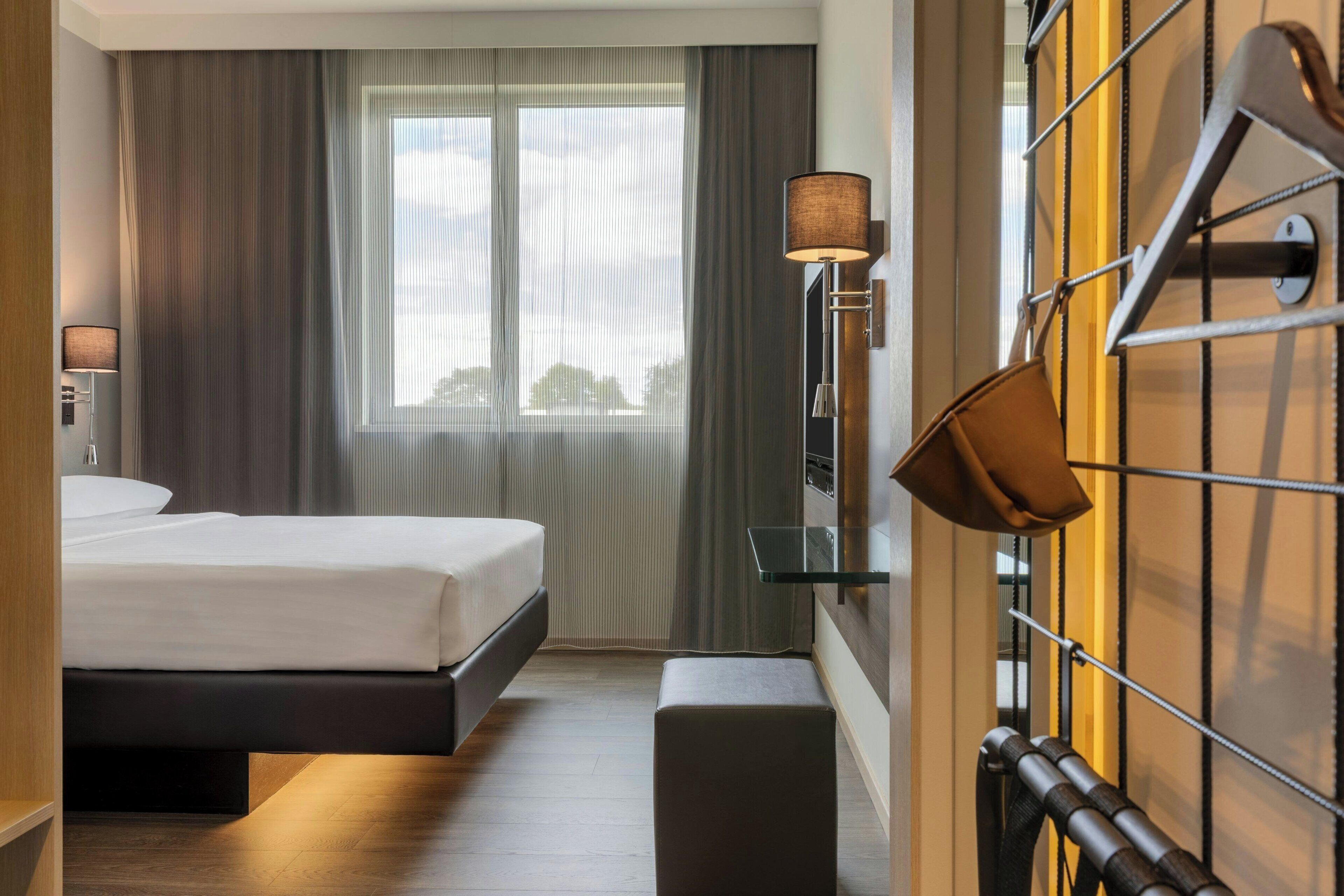 A hotel room with a bed, a nightstand, a lamp, a window with curtains, and a wall-mounted coat rack.