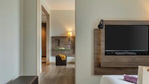 A modern hotel room with a large flat-screen TV mounted on the wall, a comfortable sofa, a small ottoman, and a bedside table with a lamp.