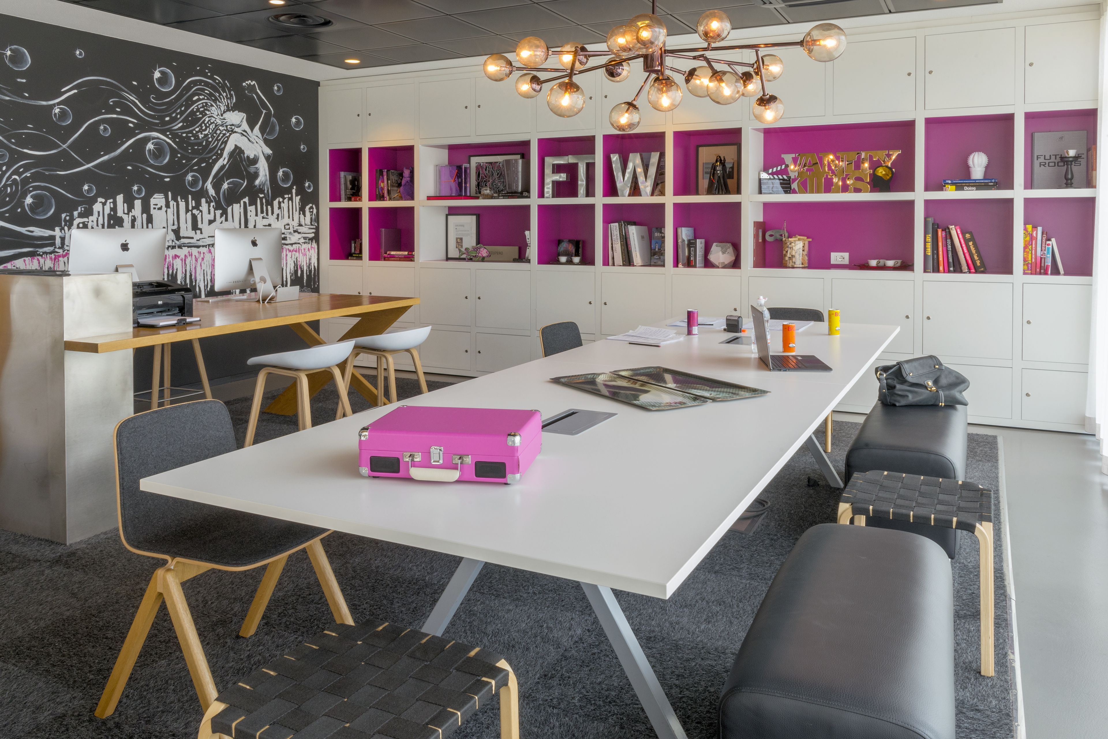 Ein moderner Arbeitsraum mit einem weißen Tisch, schwarzen und hölzernen Stühlen, einem pinken Koffer und einer schwarzen Bank.