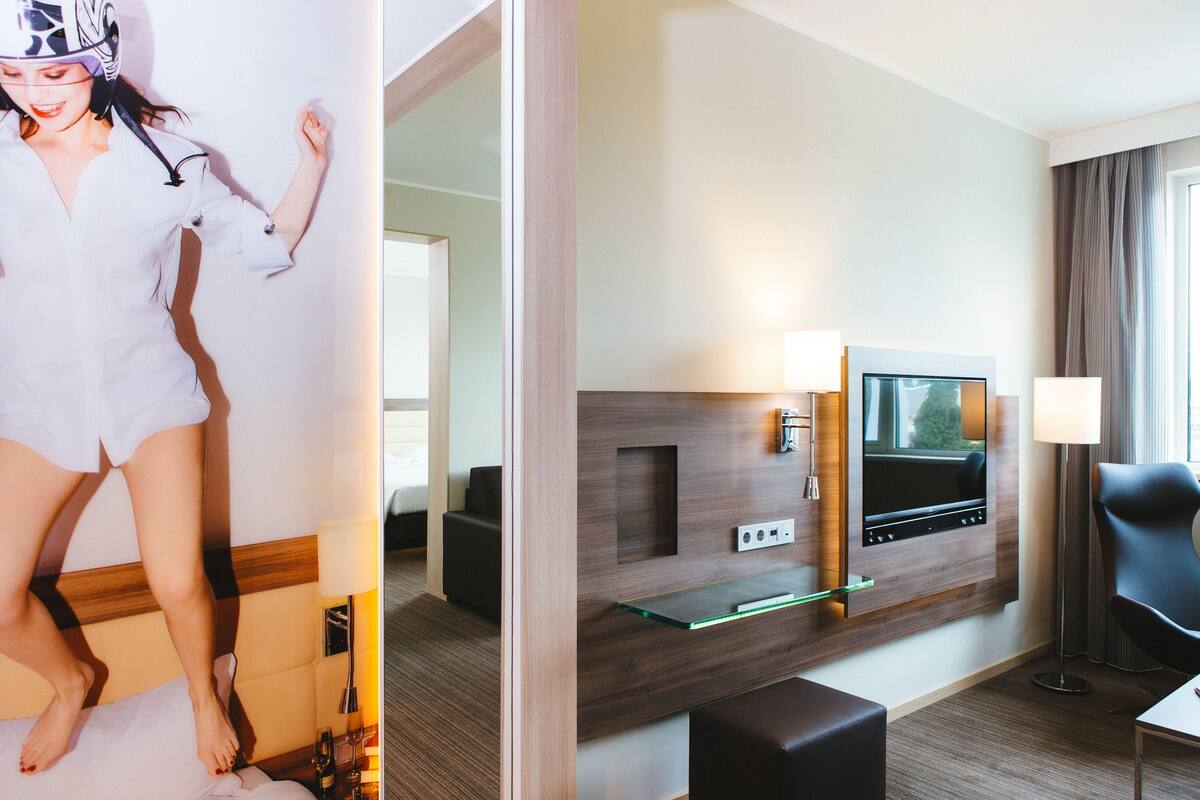 A hotel room with a wooden headboard, a flat-screen TV, a glass shelf, a black leather chair, and a bedside table with a lamp.