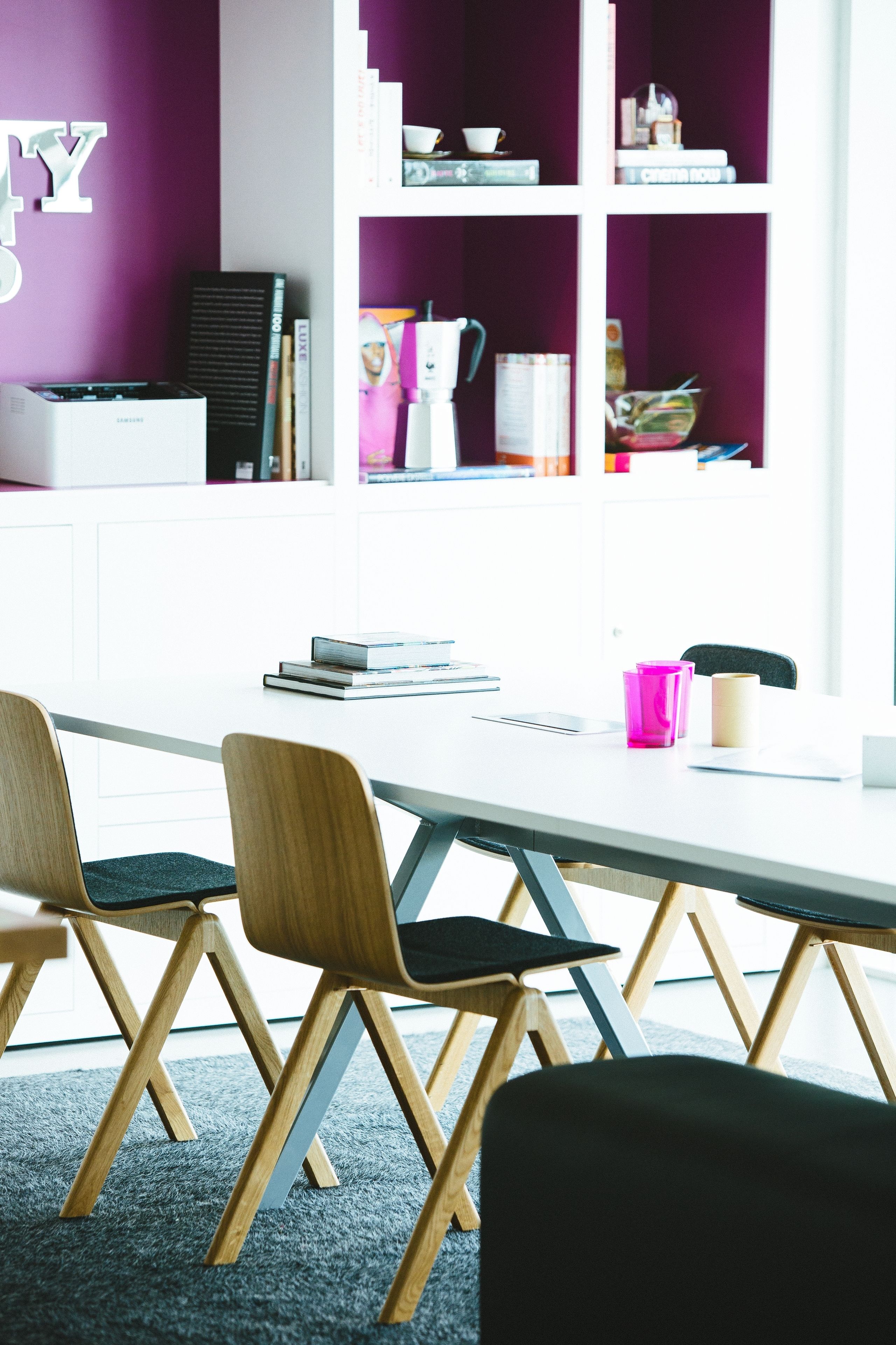 A modern meeting room with a white table, wooden chairs, and a bookshelf with various items.