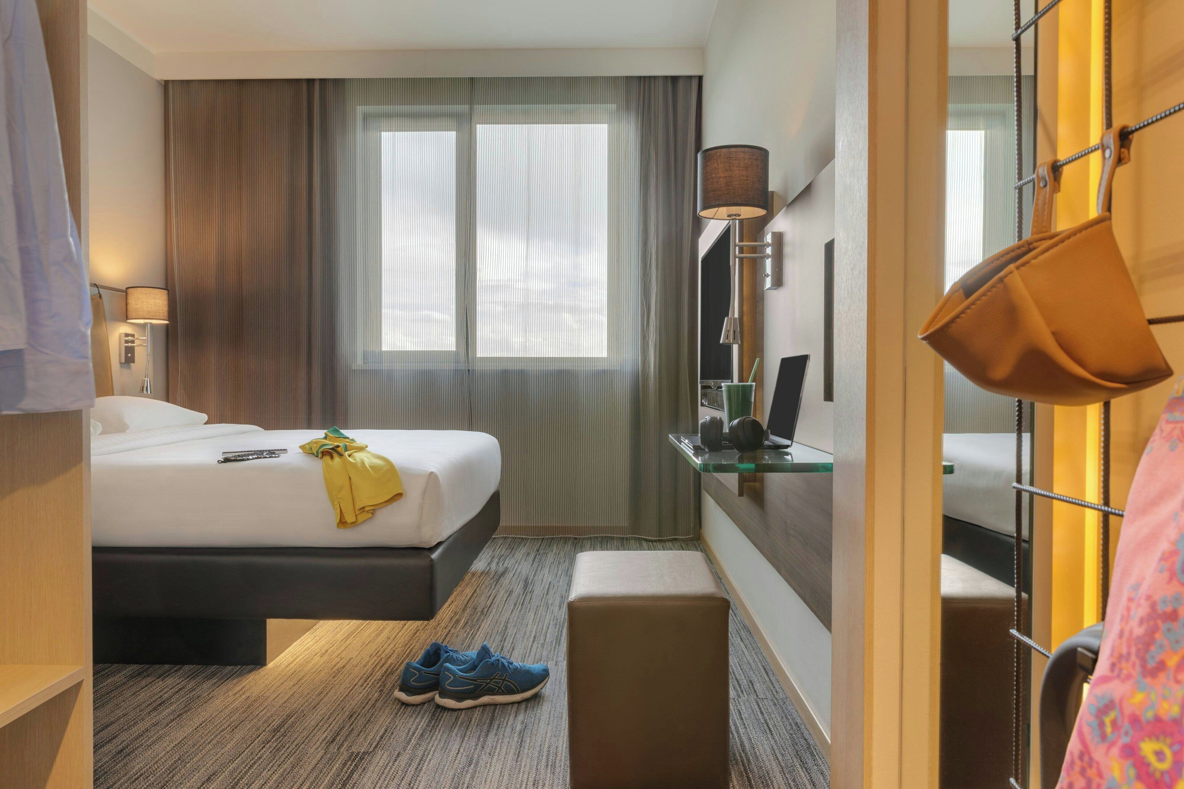 A hotel room with a bed, bedside table, lamp, and a window with curtains.