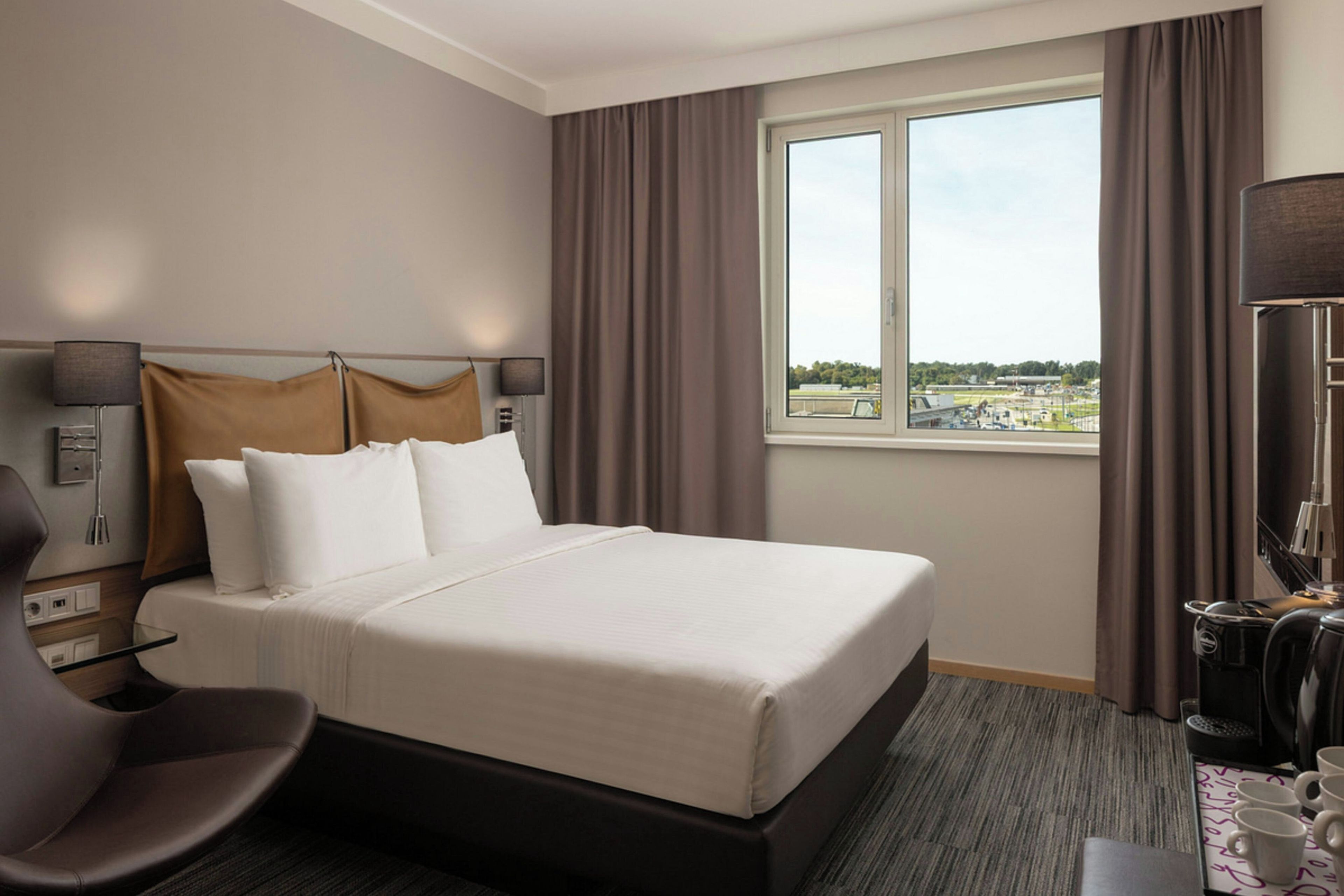 A hotel room with a large bed, a desk, a chair, a window with a view, and a lamp.