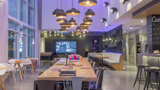 A modern dining area with a long wooden table, multiple chairs, hanging pendant lights, and a projection screen in the background.