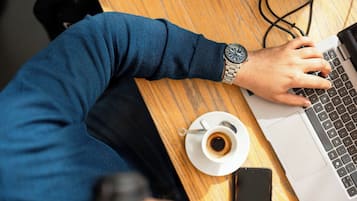 Una persona utilitzant un portàtil sobre una taula de fusta, amb una tassa de cafè i un telèfon mòbil a prop.